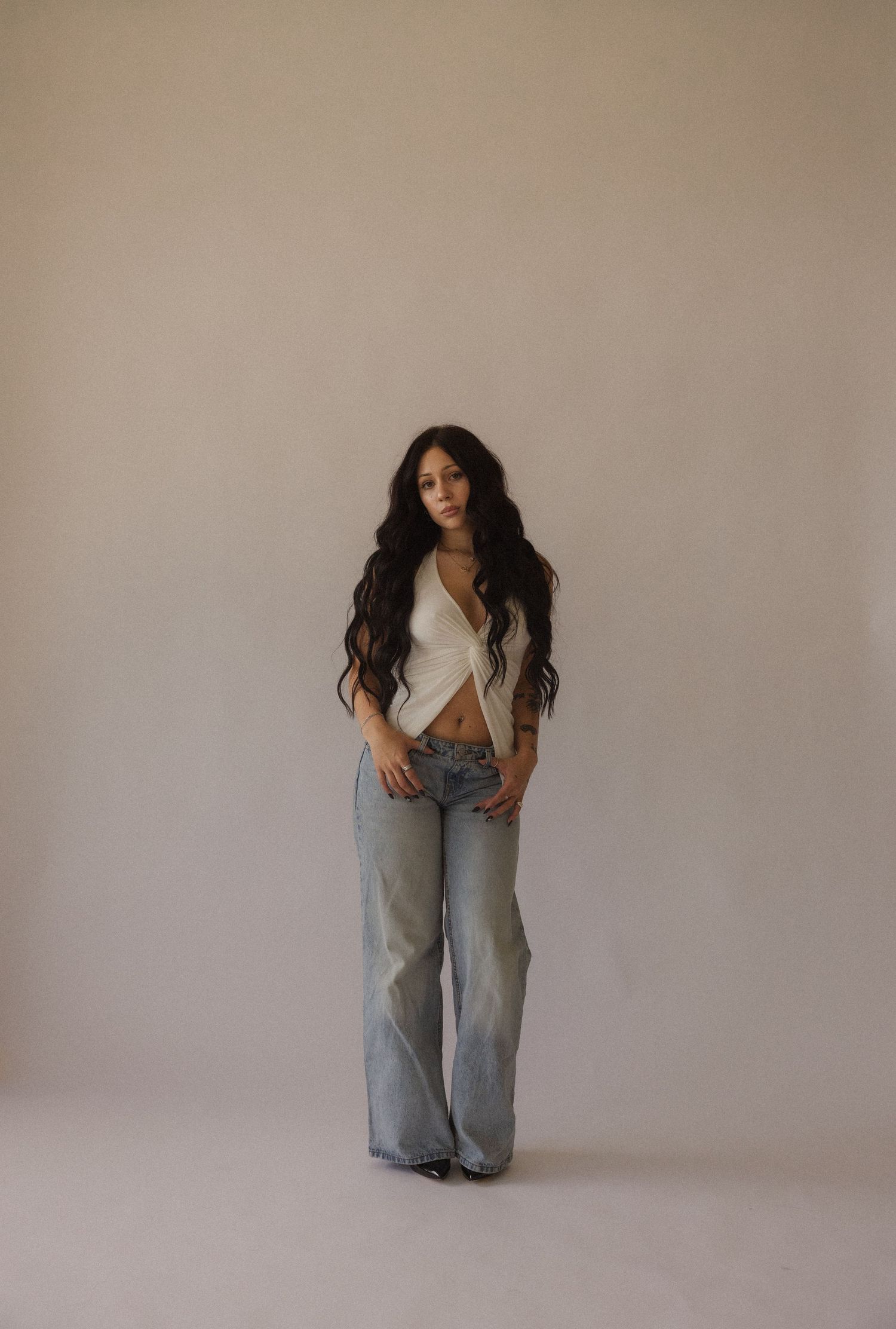 Woman with long wavy black hair wearing a white crop top and light wash wide-leg jeans, posing against a neutral wall.