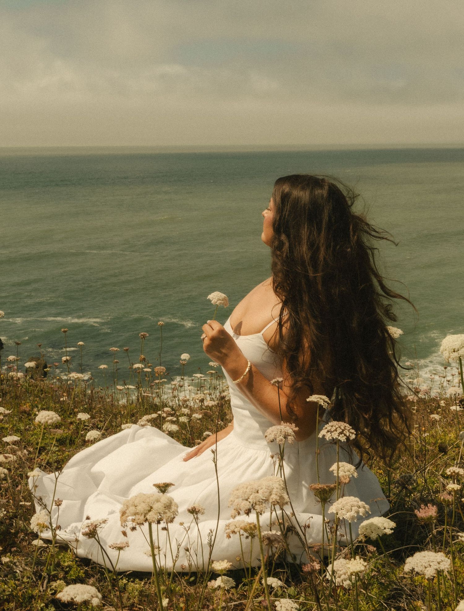 Woman in white dress sitting among wildflowers on coastal cliffside, long dark hair blowing in wind with ocean backdrop.