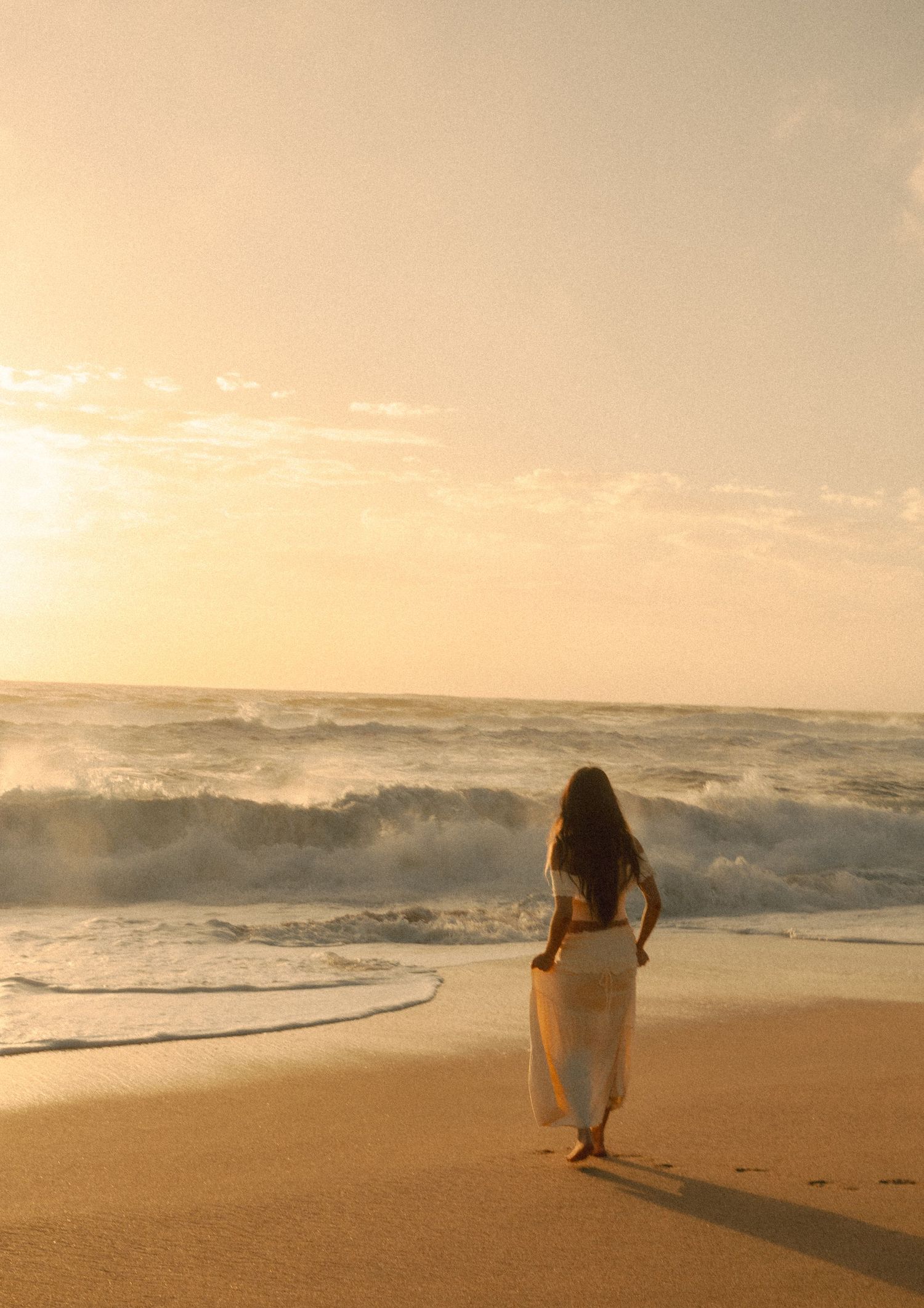 Woman in white skirt walking on sandy beach toward crashing ocean waves at golden sunset.