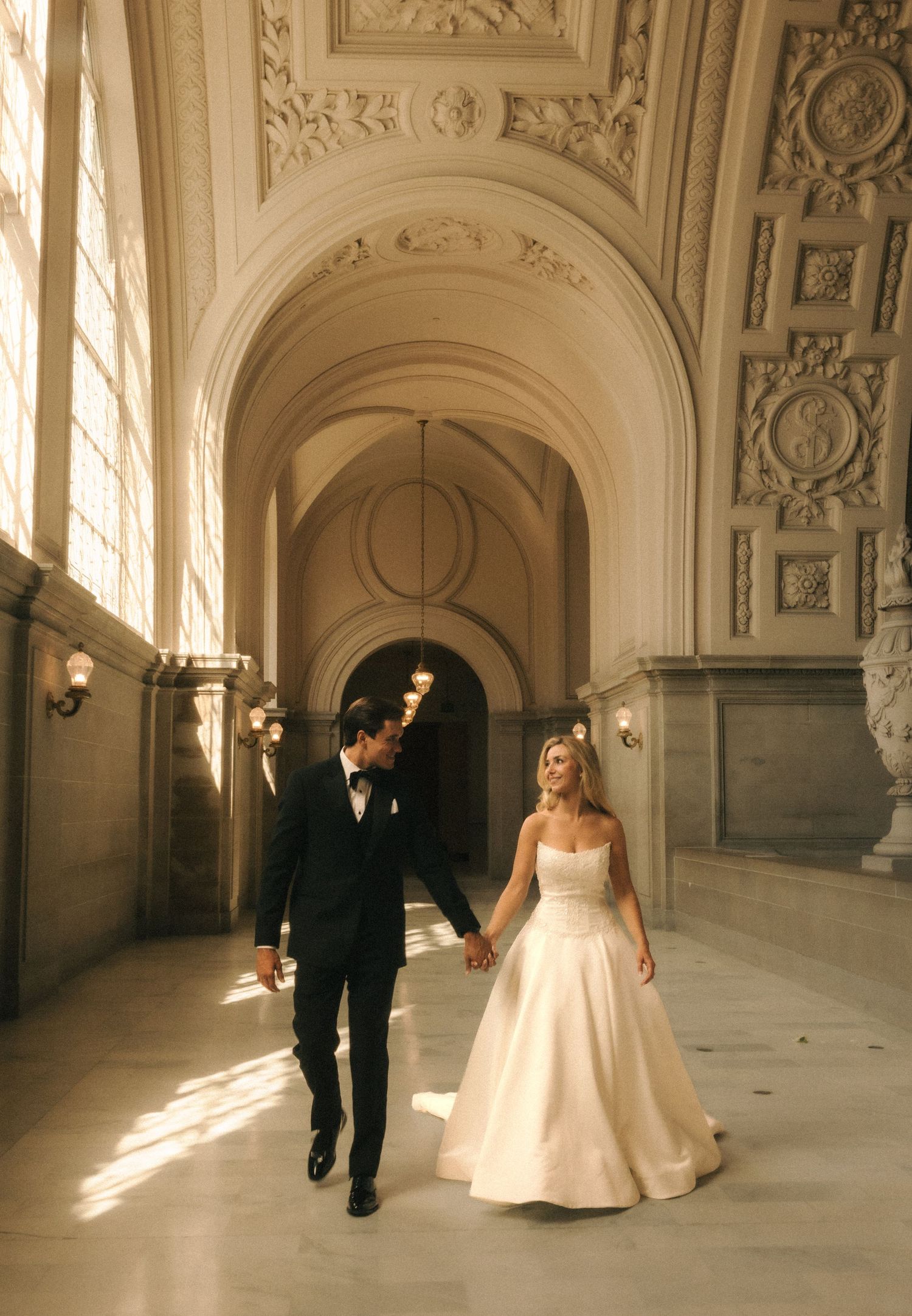 Bride and groom walk hand in hand through ornate grand hall with arched ceilings and marble floors.