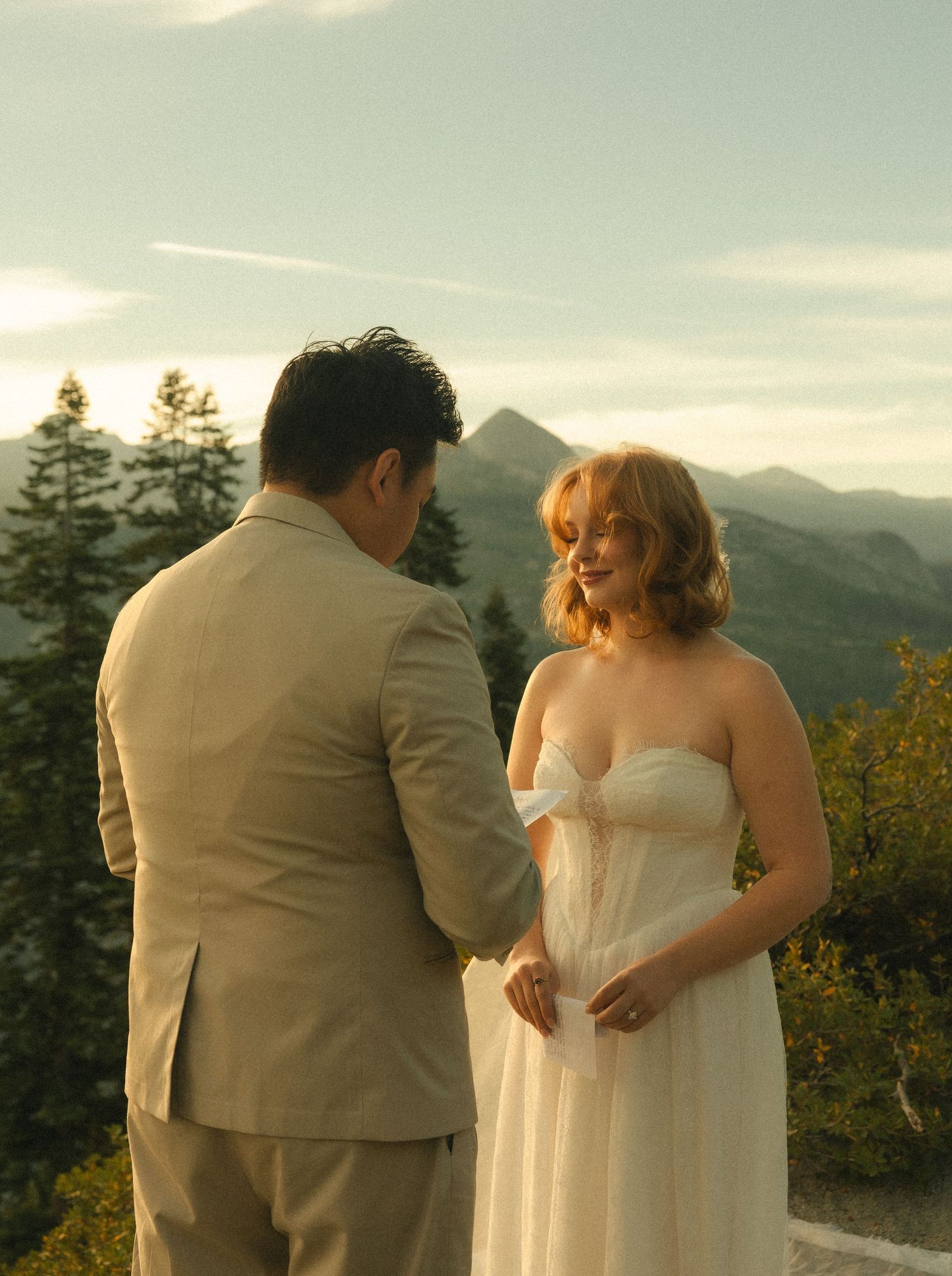 Couple in wedding attire sharing a romantic moment outdoors with mountain and forest backdrop at golden hour.