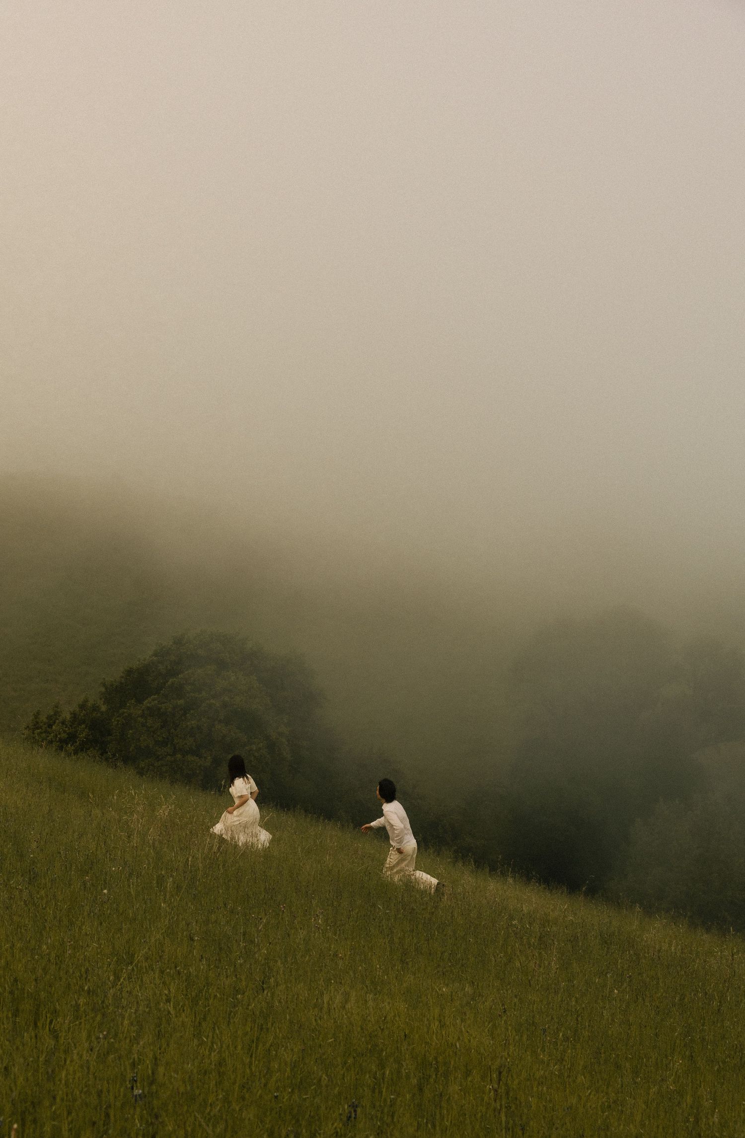 Two people in white clothing walk through a misty green hillside with fog-covered trees in the background.