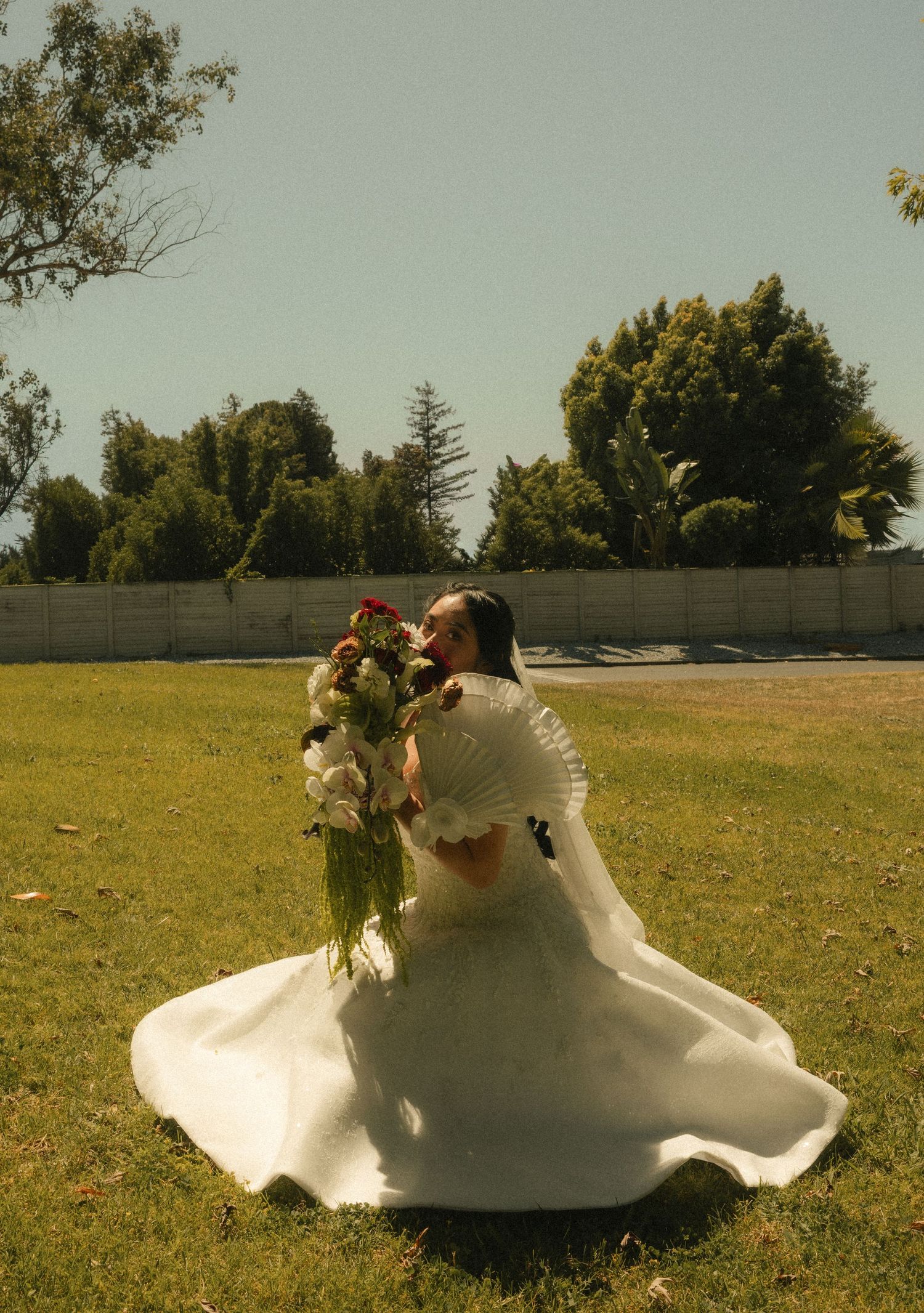 Bride in white gown twirling on green lawn, holding floral bouquet, with trees and wall in background.
