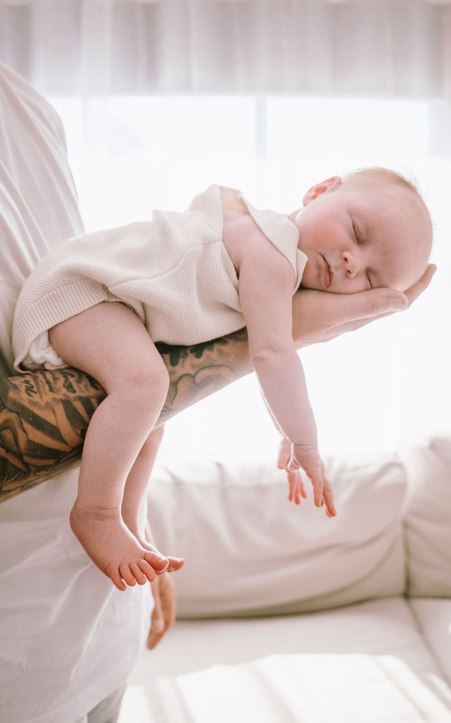 A sleeping baby in a white onesie rests peacefully on a bright white couch in soft natural light.