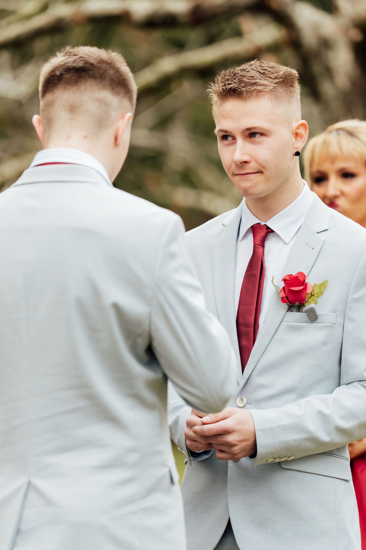 Two grooms in gray suits with red ties hold hands during an outdoor wedding ceremony.