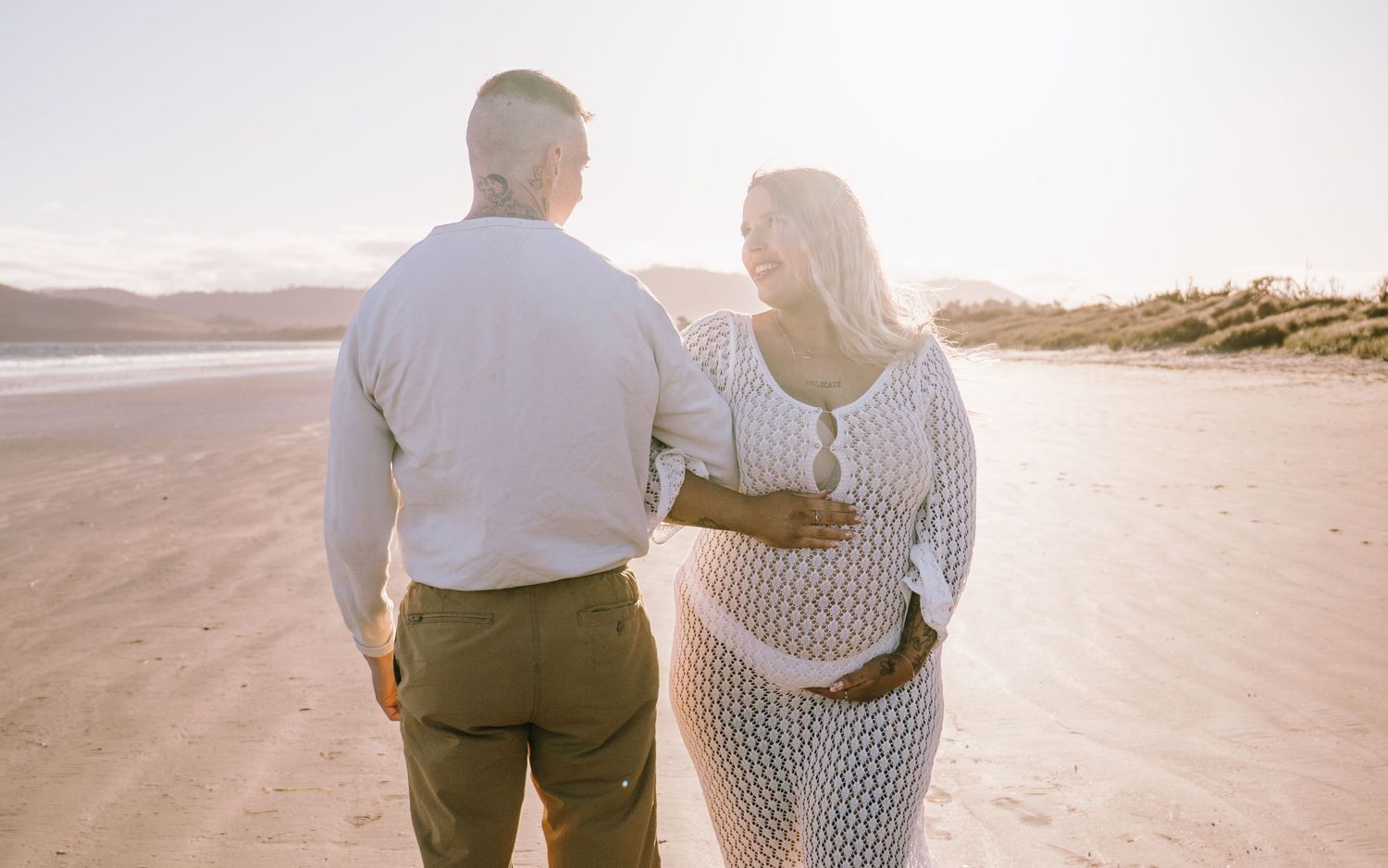 Couple walking together on a sandy beach at sunset holding each other close in romantic embrace.