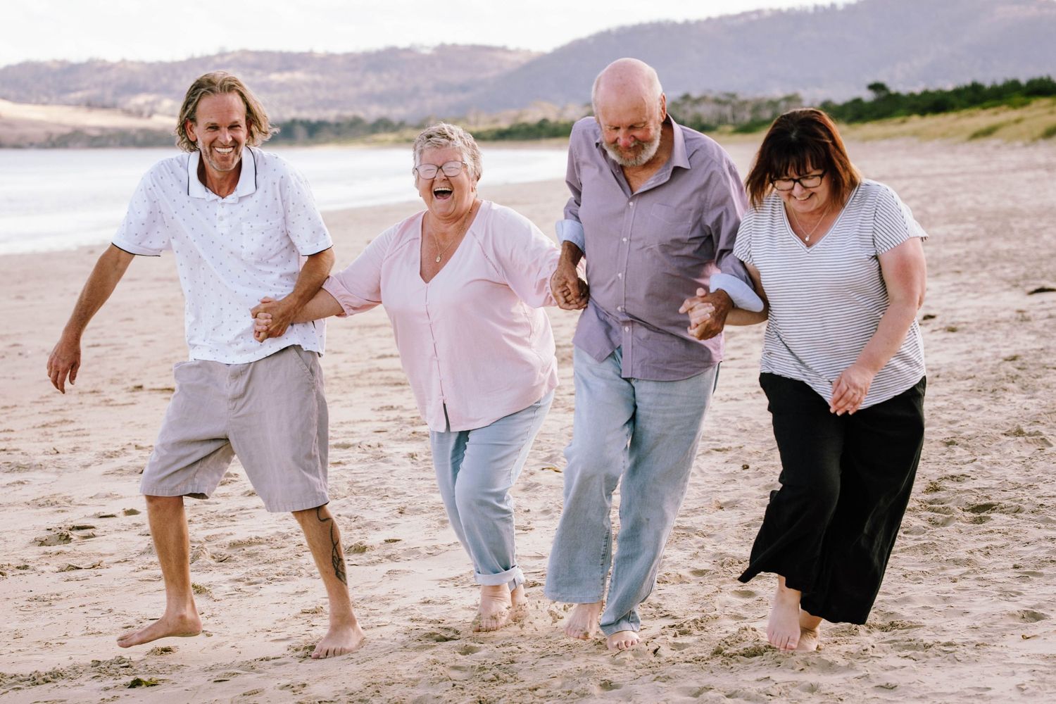 A group of four people laughing and walking together on a sandy beach with mountains in the background.