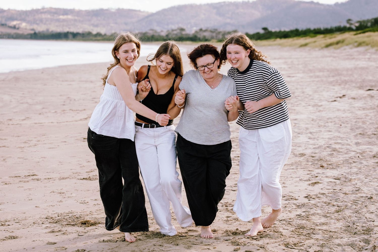 A group of friends laugh together while walking along a scenic beach with mountains in the background.