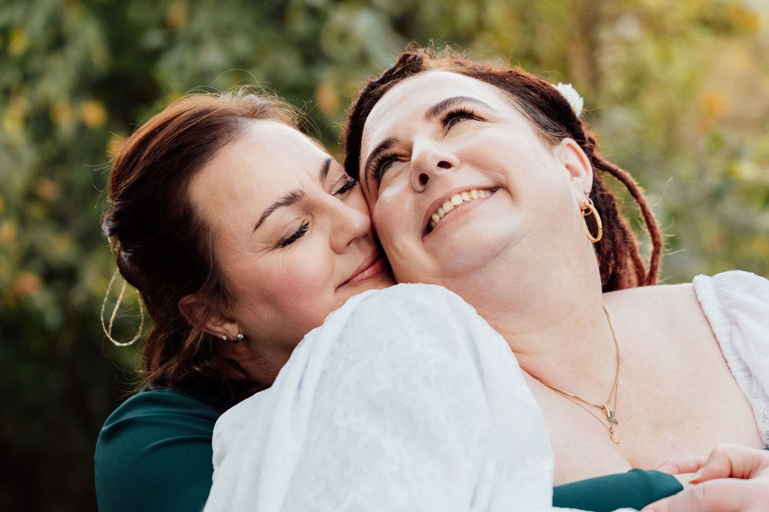 Two women share a tender moment outdoors, one kissing the other's cheek as she smiles warmly in a white dress.