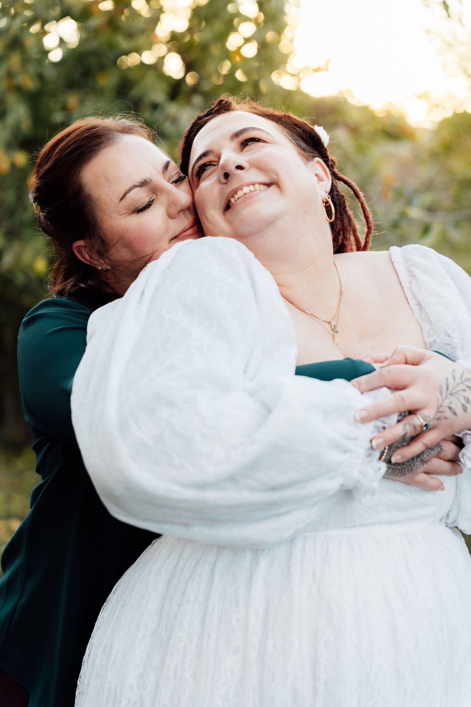 Two LQBTQ+ women embrace outdoors at golden hour, one in white bridal gown, other in teal button down shirt, sharing a tender moment.