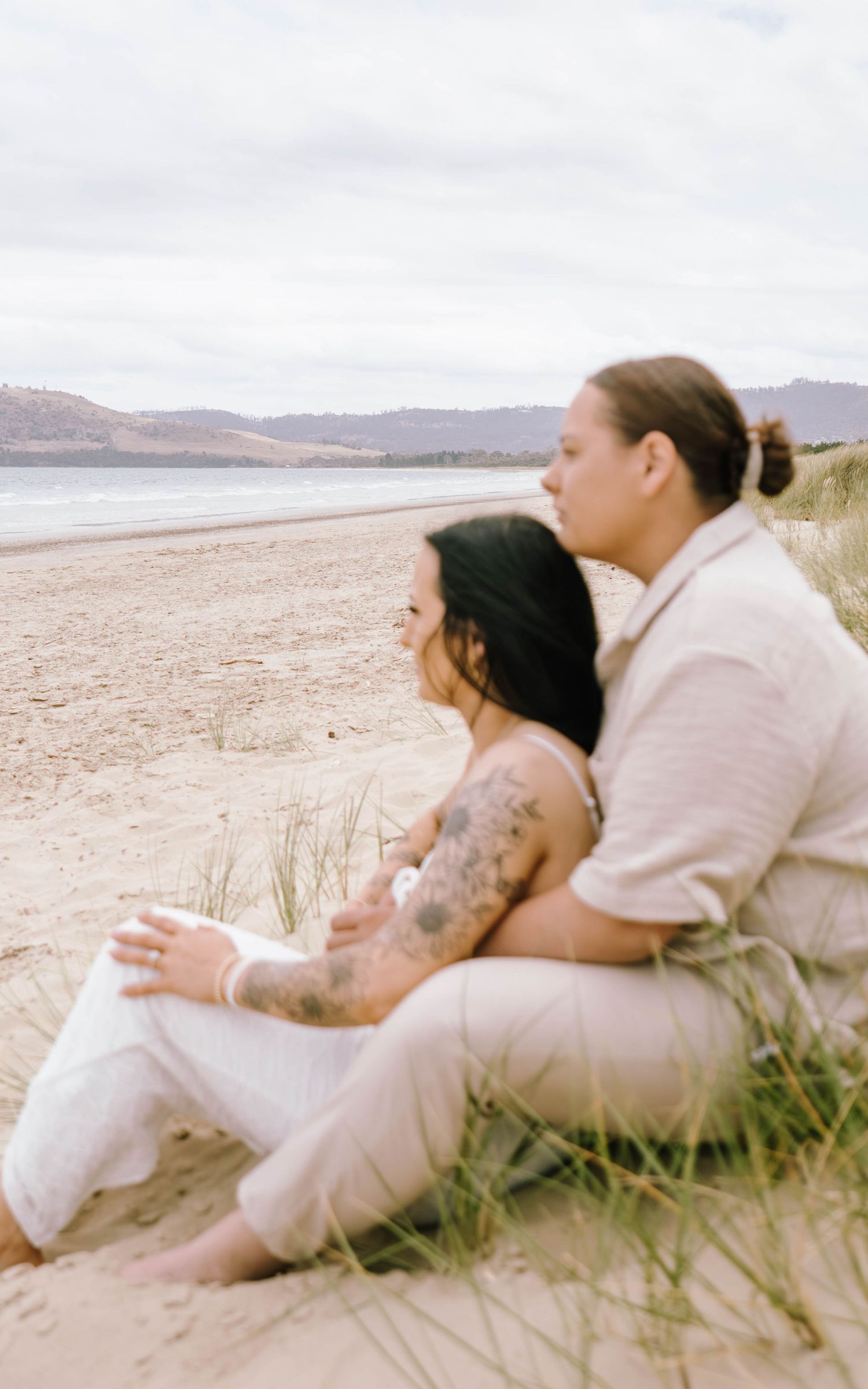 Two people sitting together on coastal dunes overlooking a beach landscape.