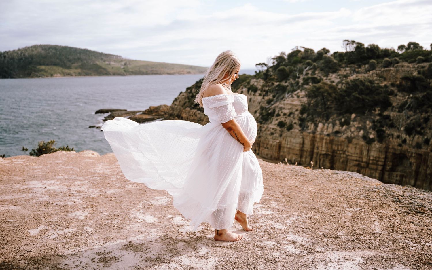A white flowing dress catches the wind on a dramatic clifftop overlooking a serene lake and tree-lined shore.