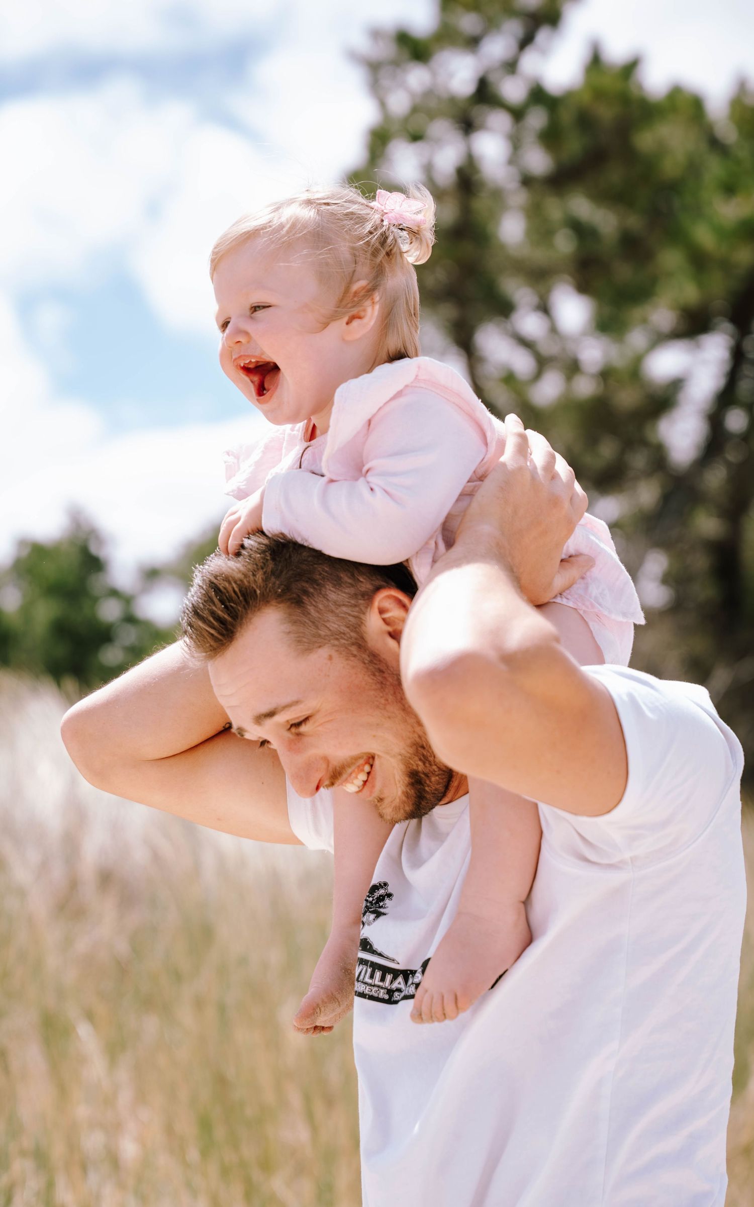 A playful outdoor scene with a laughing baby in pink riding on shoulders against a blue sky and greenery.