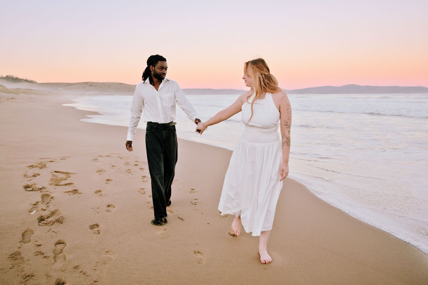 Interracial couple walking hand-in-hand on sandy beach at sunset, woman in white dress, man in white shirt and black pants.