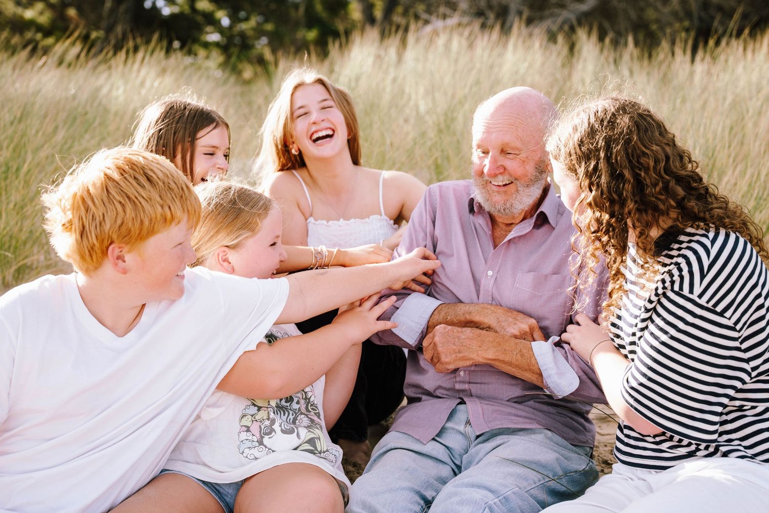 Multi-generational family laughing together outdoors on a sunny day, sitting in a grassy field during a joyful gathering.
