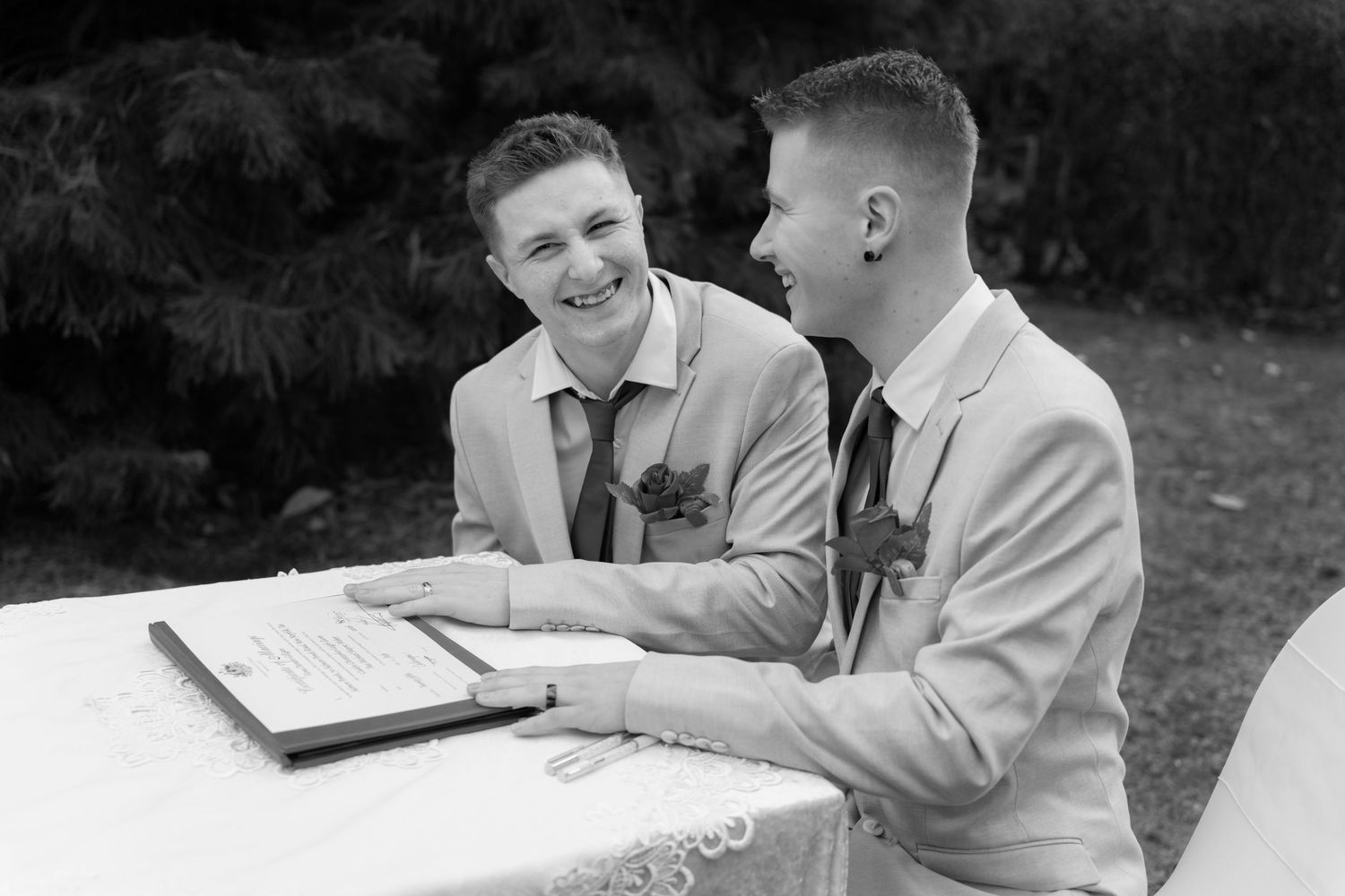 Two grooms laughing and signing wedding certificate outdoors in black and white photo.