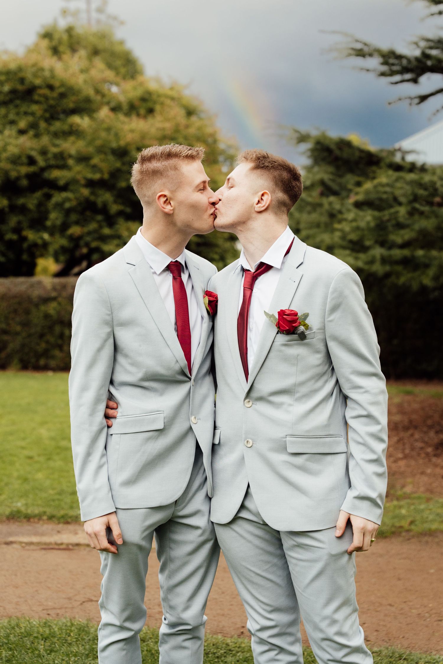 Two grooms in matching light gray suits with red ties share a kiss outdoors with a rainbow in the background.