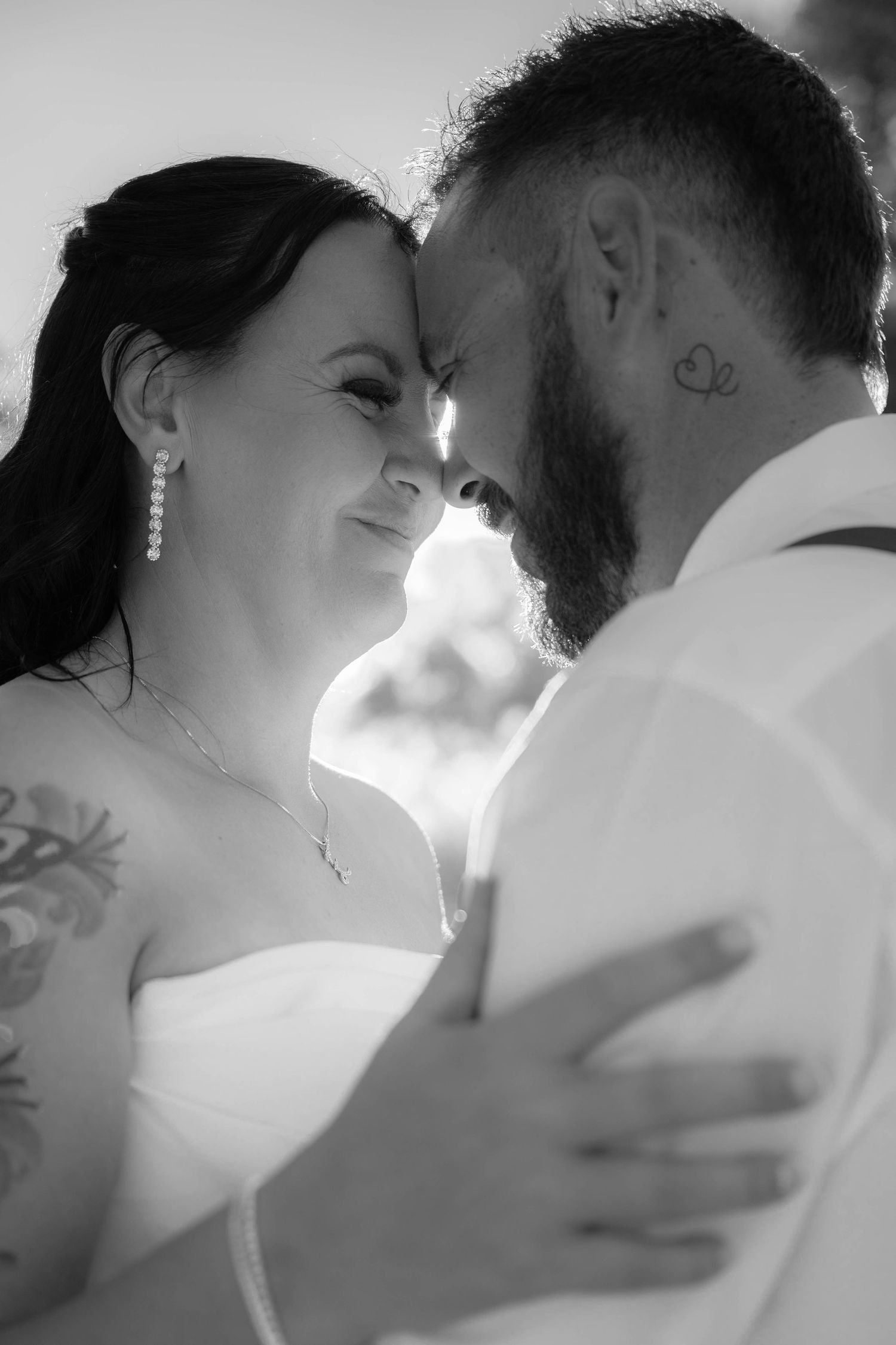 Black and white wedding photo of bride and groom touching foreheads, sharing an intimate moment, both with tattoos visible.