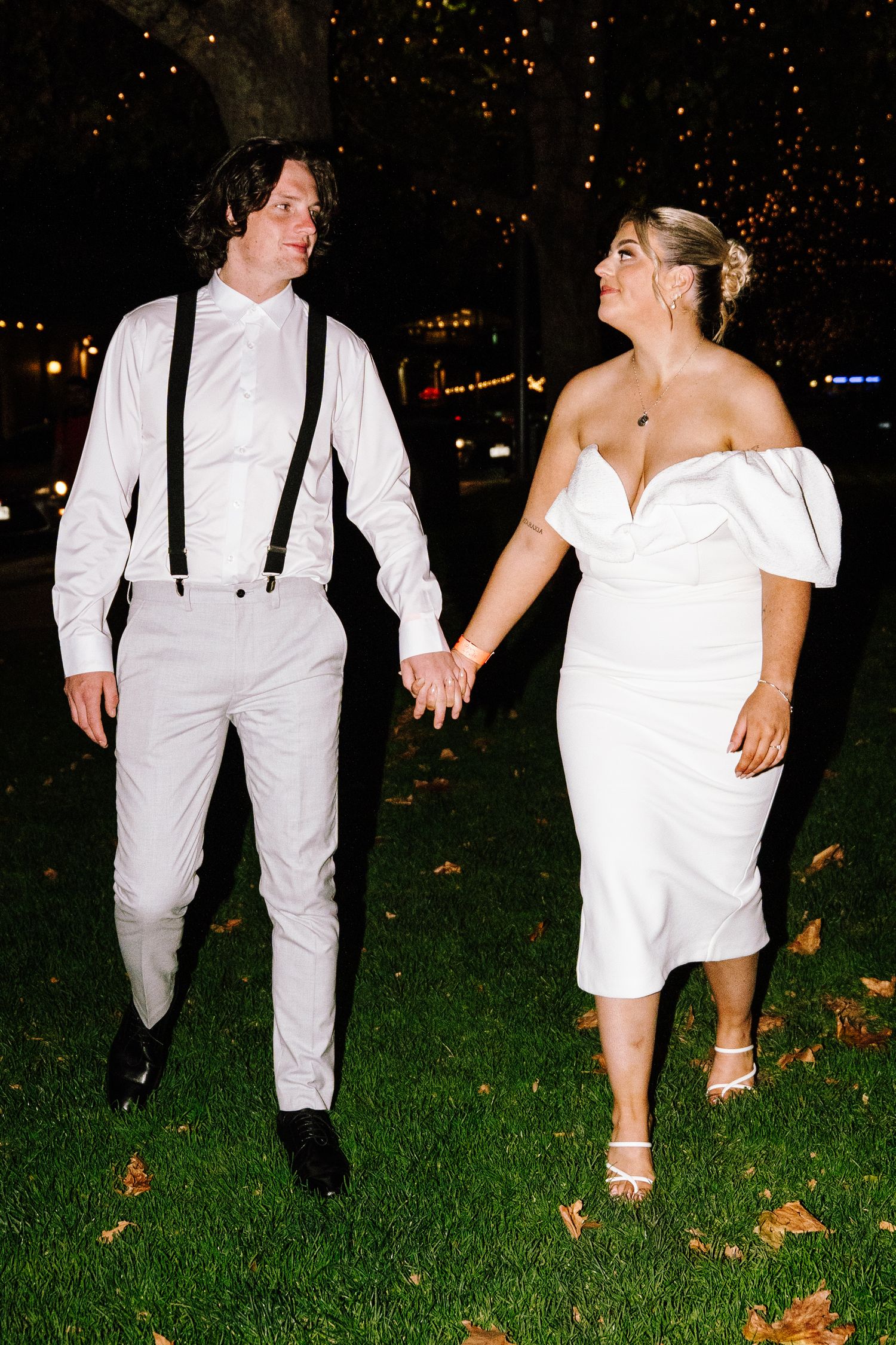 Bride and groom holding hands walking on grass at night under fairy lights.