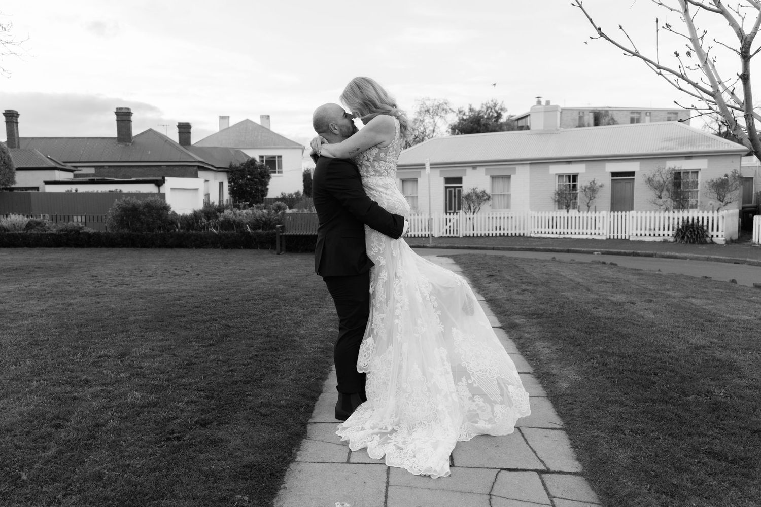 Black and white photo of groom lifting bride in wedding dress on garden path outside elegant venue.