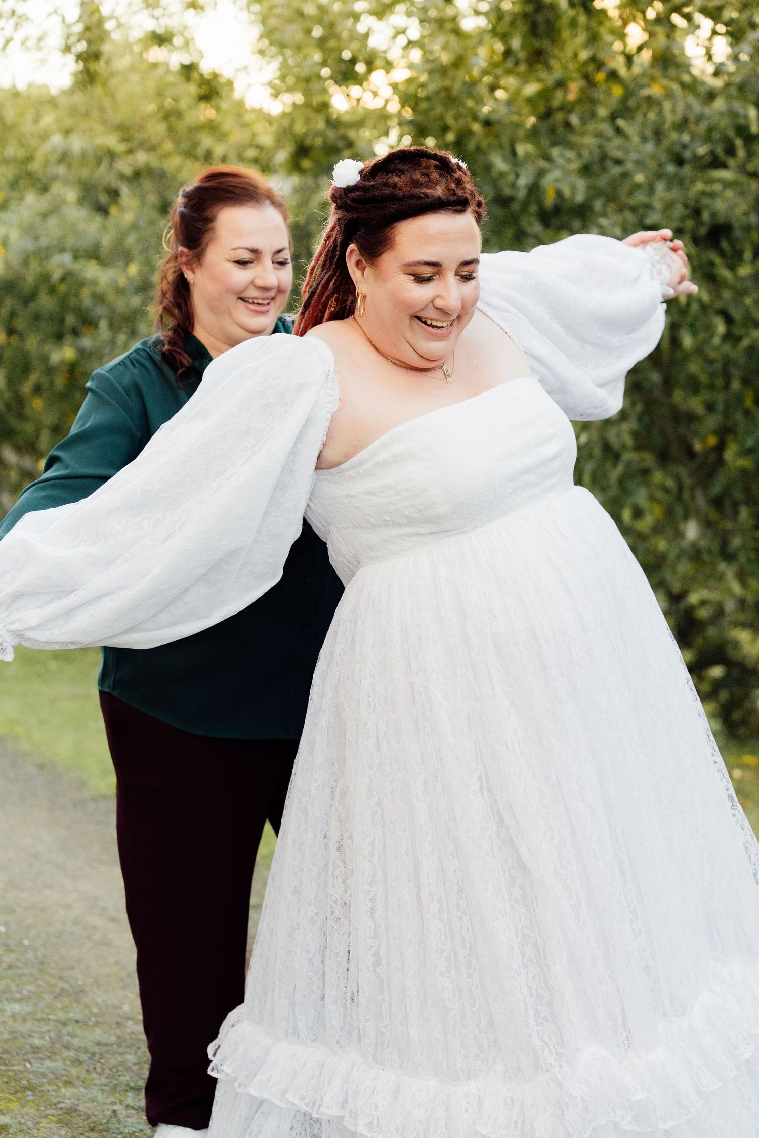 A bride in a white lace gown with puff sleeves laughs as the other bride in green move like aeroplanes outdoors.