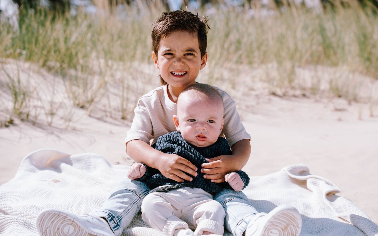 Two children sitting together on a blanket at a sandy beach on a sunny day.