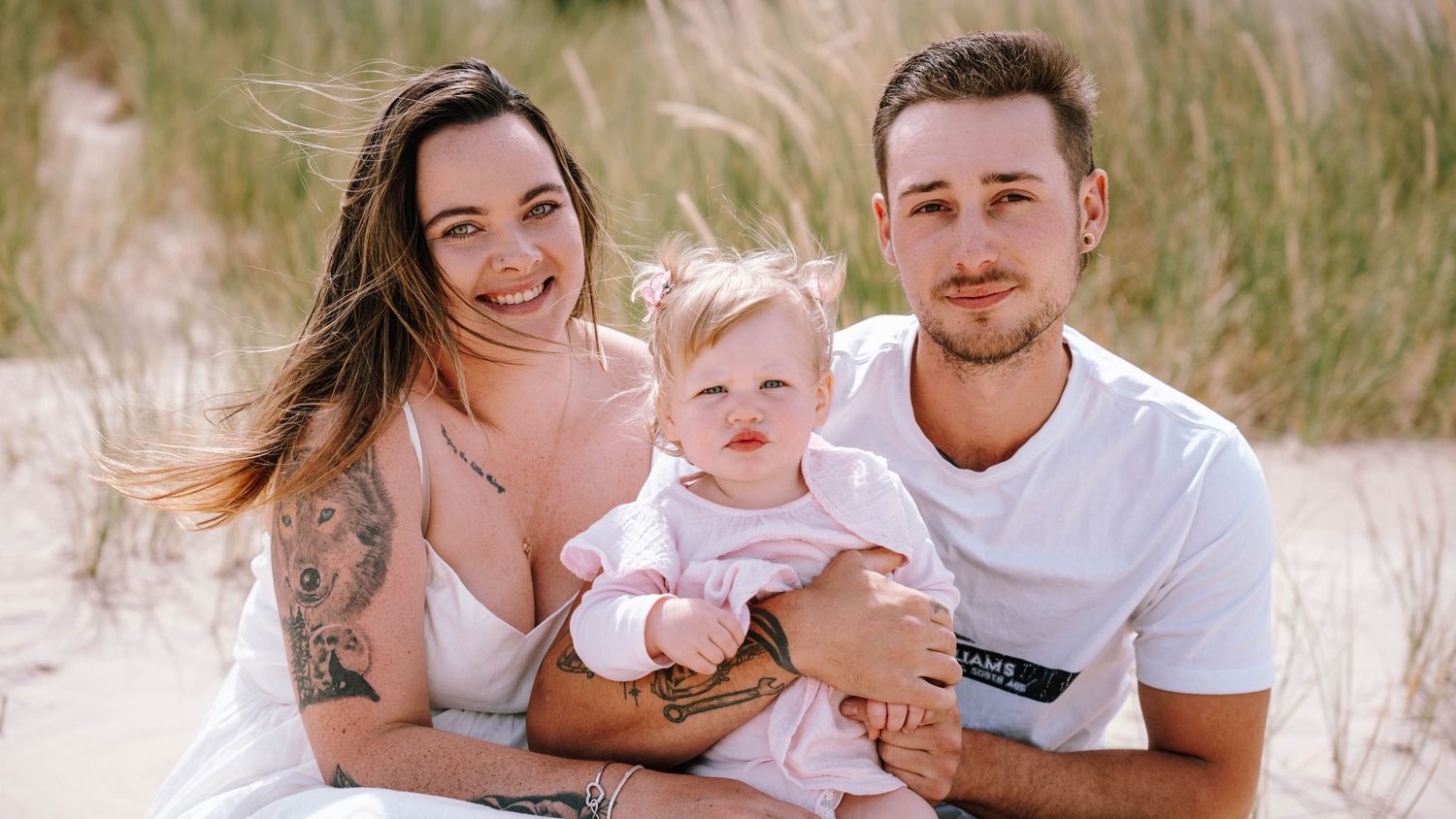 Young family with tattooed mother, father in white tee, and baby girl posing together on a sandy beach with dune grass.