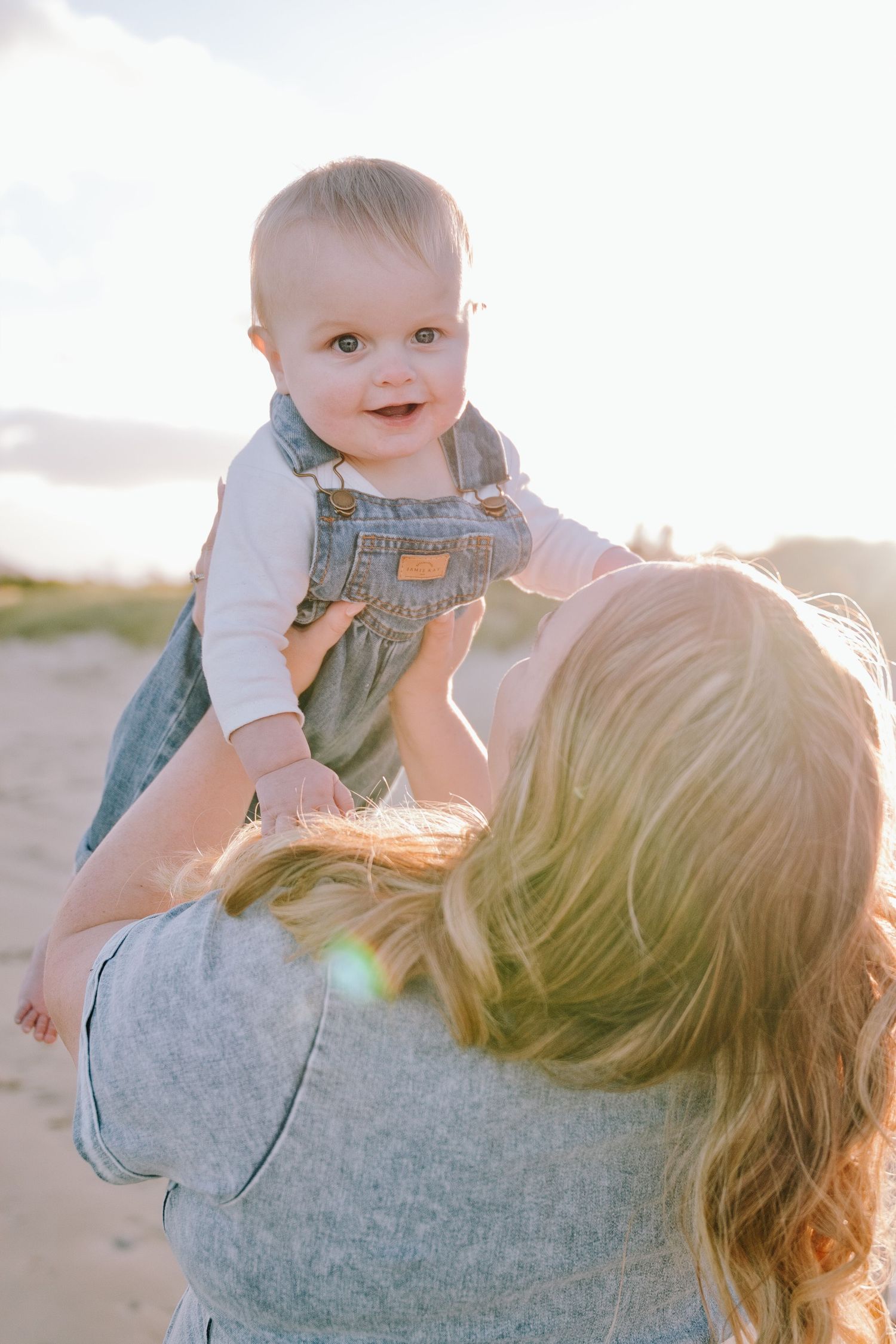 Mother lifting smiling baby in denim overalls against a golden sunset sky outdoors.