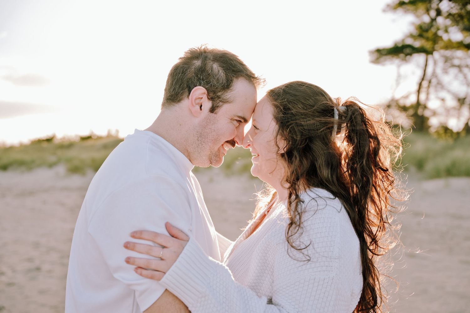 A couple shares a romantic moment on a sandy beach, touching foreheads and smiling, dressed in white sweaters at golden hour.