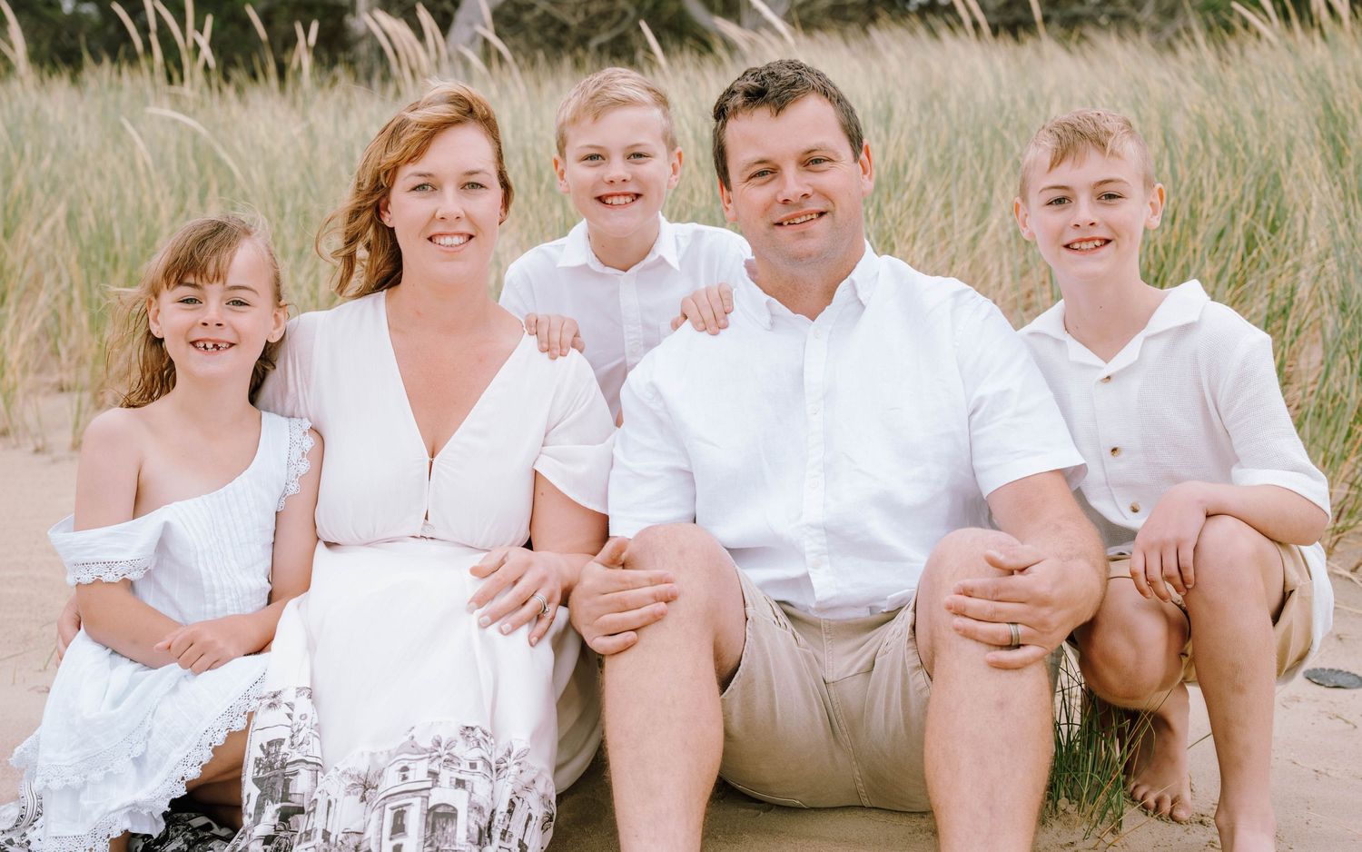 A group in white clothing sits together on grass during an outdoor photo session in warm natural lighting.
