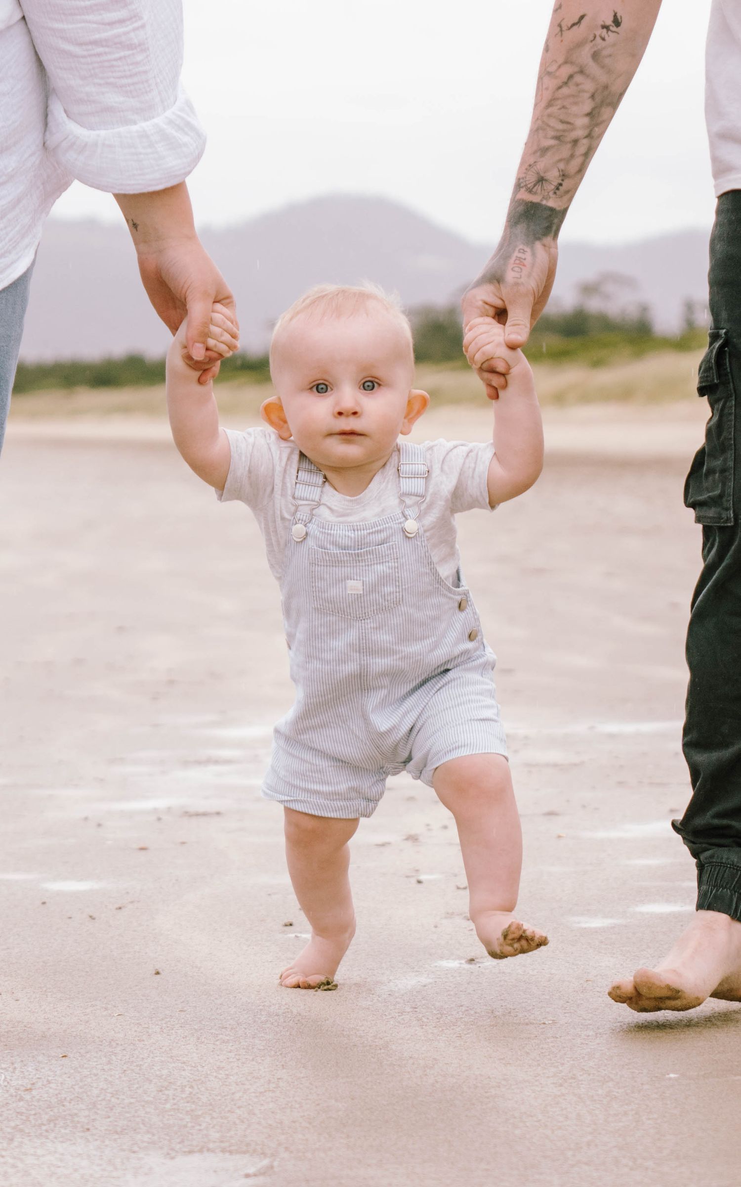 Baby in white outfit takes first steps while holding hands with adults on a sandy path.