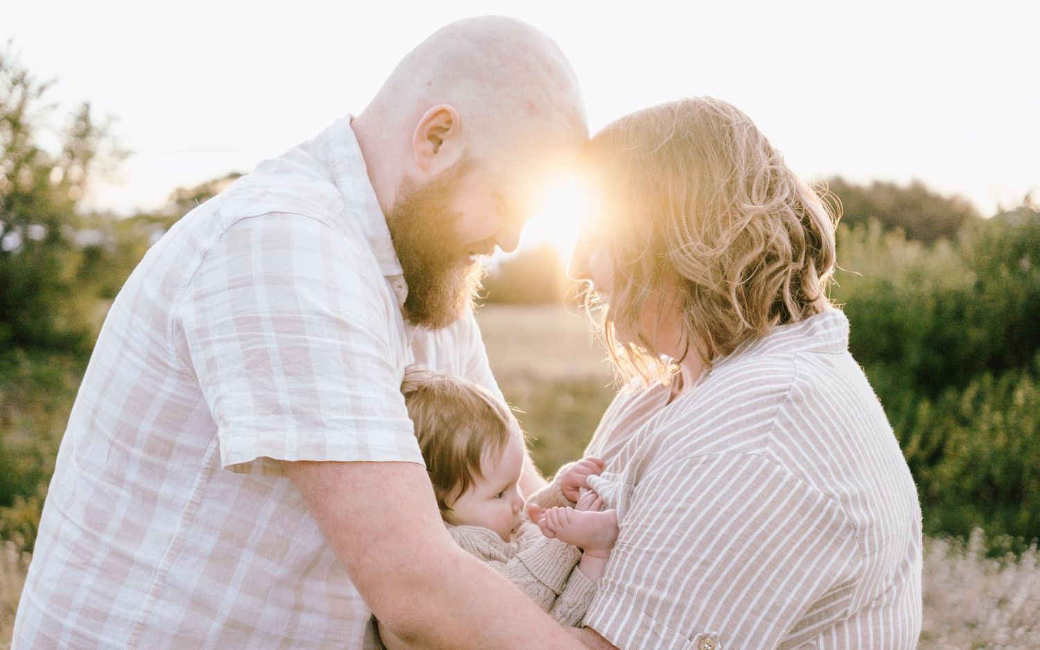 A couple tenderly holds their baby outdoors at golden hour, foreheads touching in a warm family portrait.