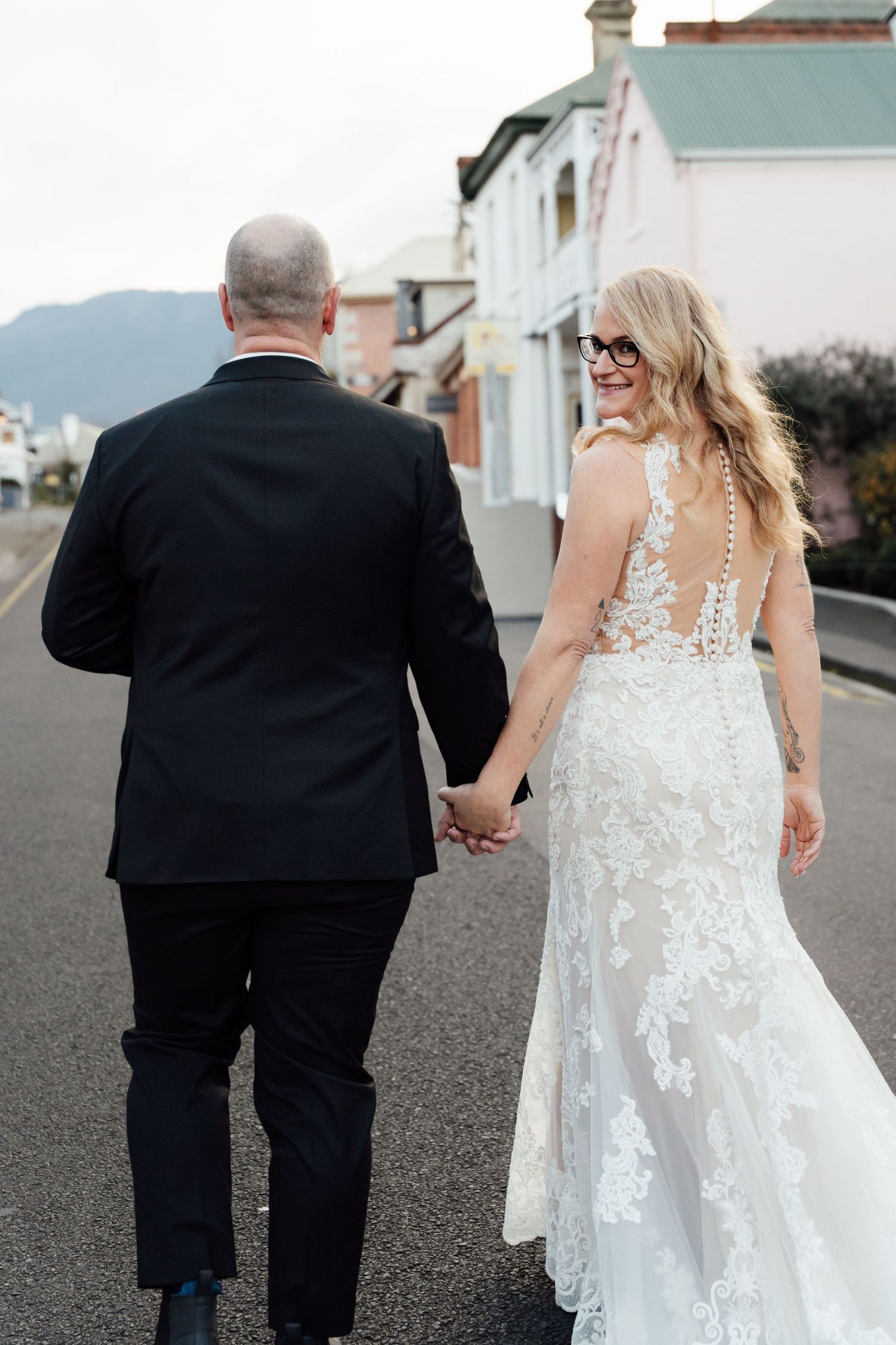A bride in lace gown and groom in black suit walk hand-in-hand down a street, she smiles back at the camera.