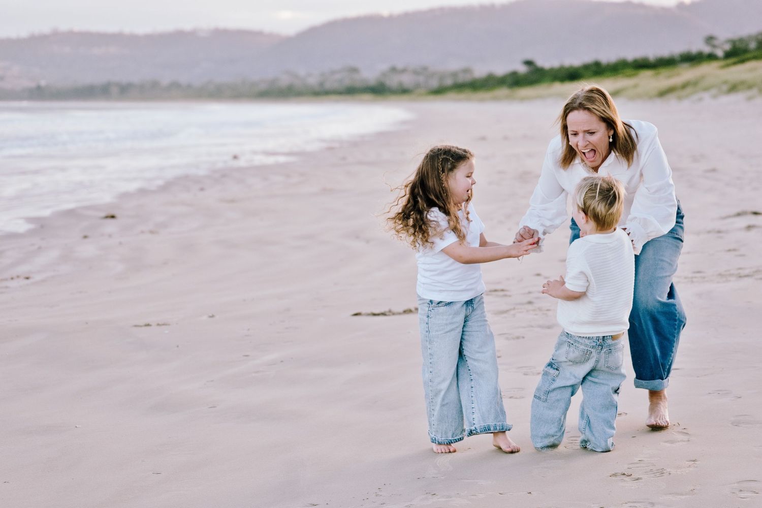 A mother plays with her two young children on a sandy beach, all wearing white tops and jeans.