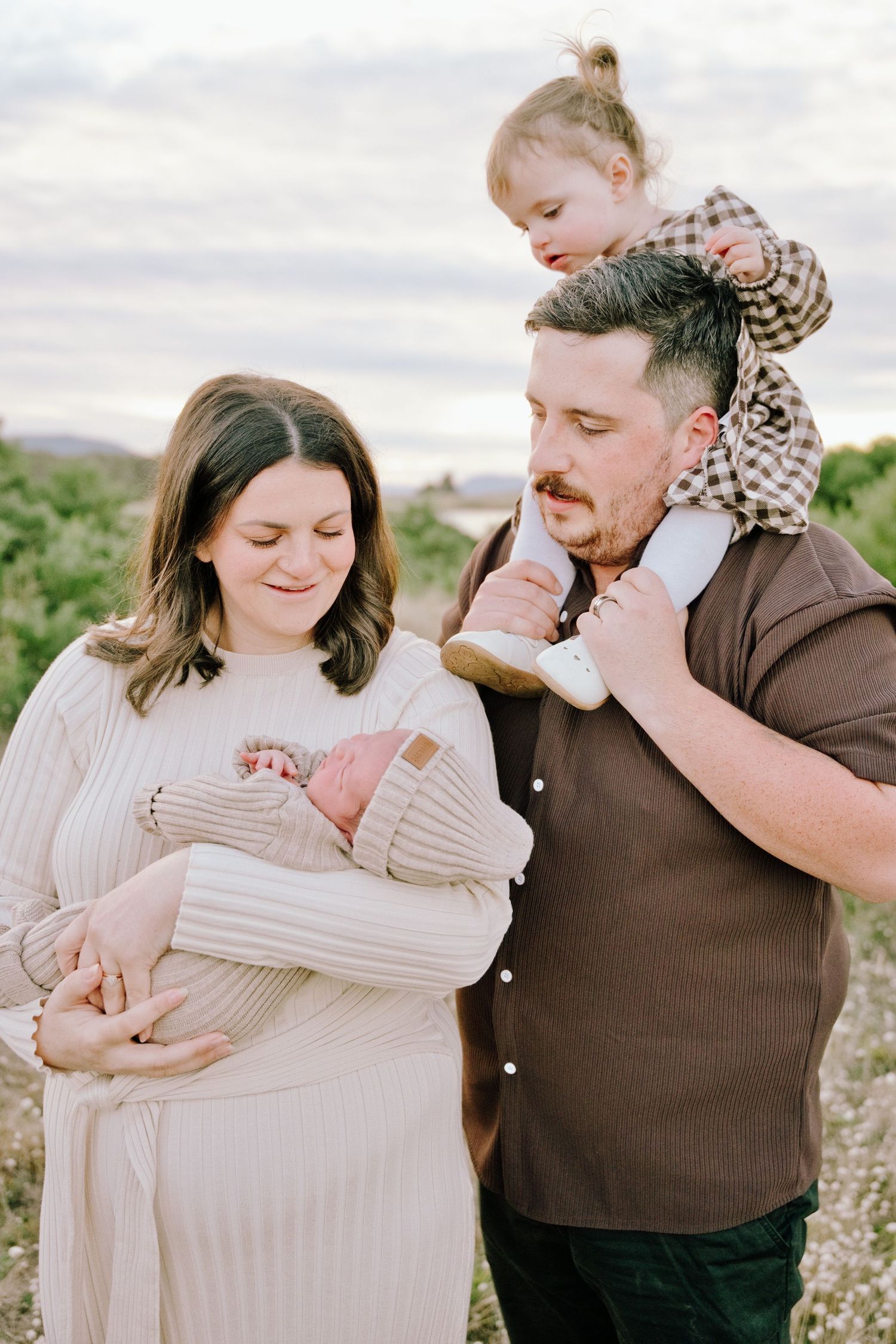 A couple stands outdoors holding their newborn baby and toddler on shoulders during a family portrait session at dusk.