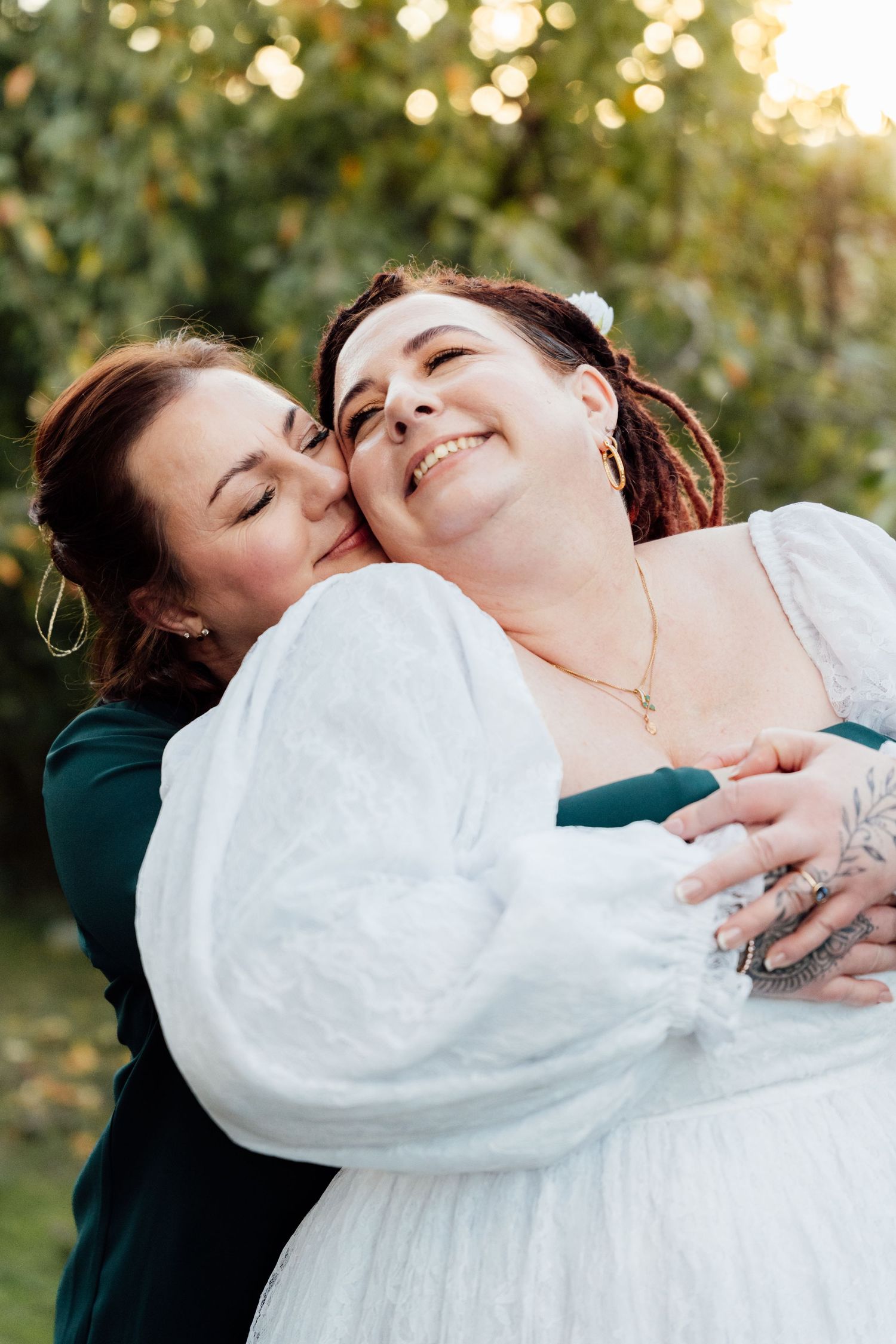 Two women share a tender embrace outdoors, one in a white bridal gown, smiling joyfully amid lush green trees.