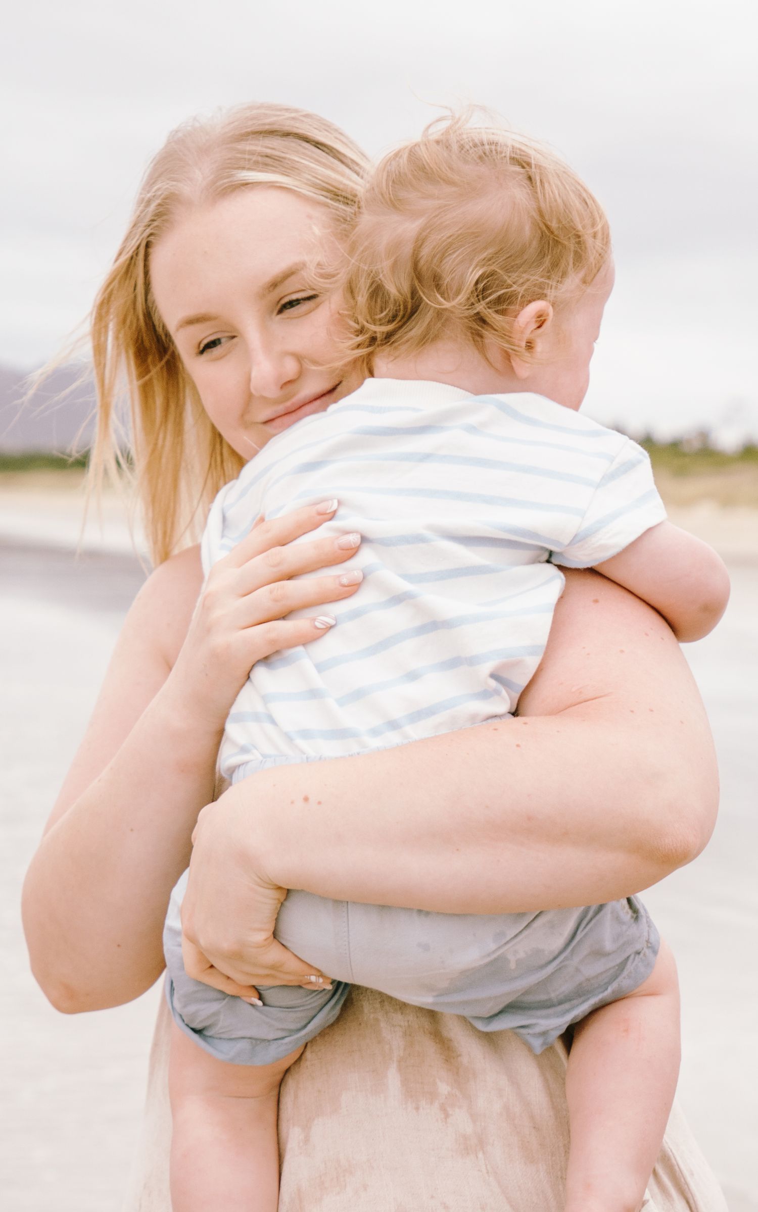 A tender moment captured as a parent embraces a baby wearing a striped outfit on a bright, sunny beach.