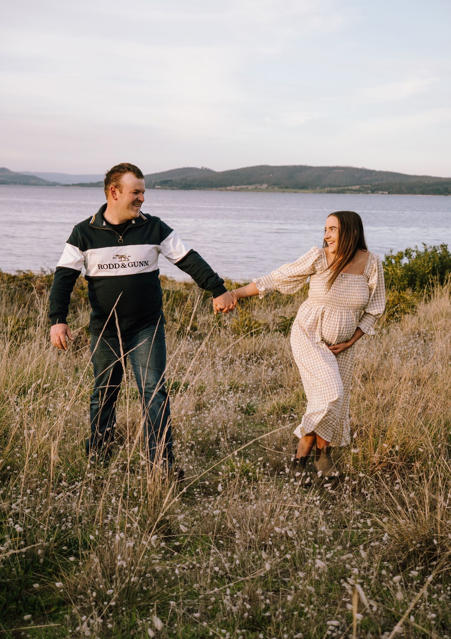 A playful couple dances and laughs together in tall grass with a scenic waterfront view in the background.