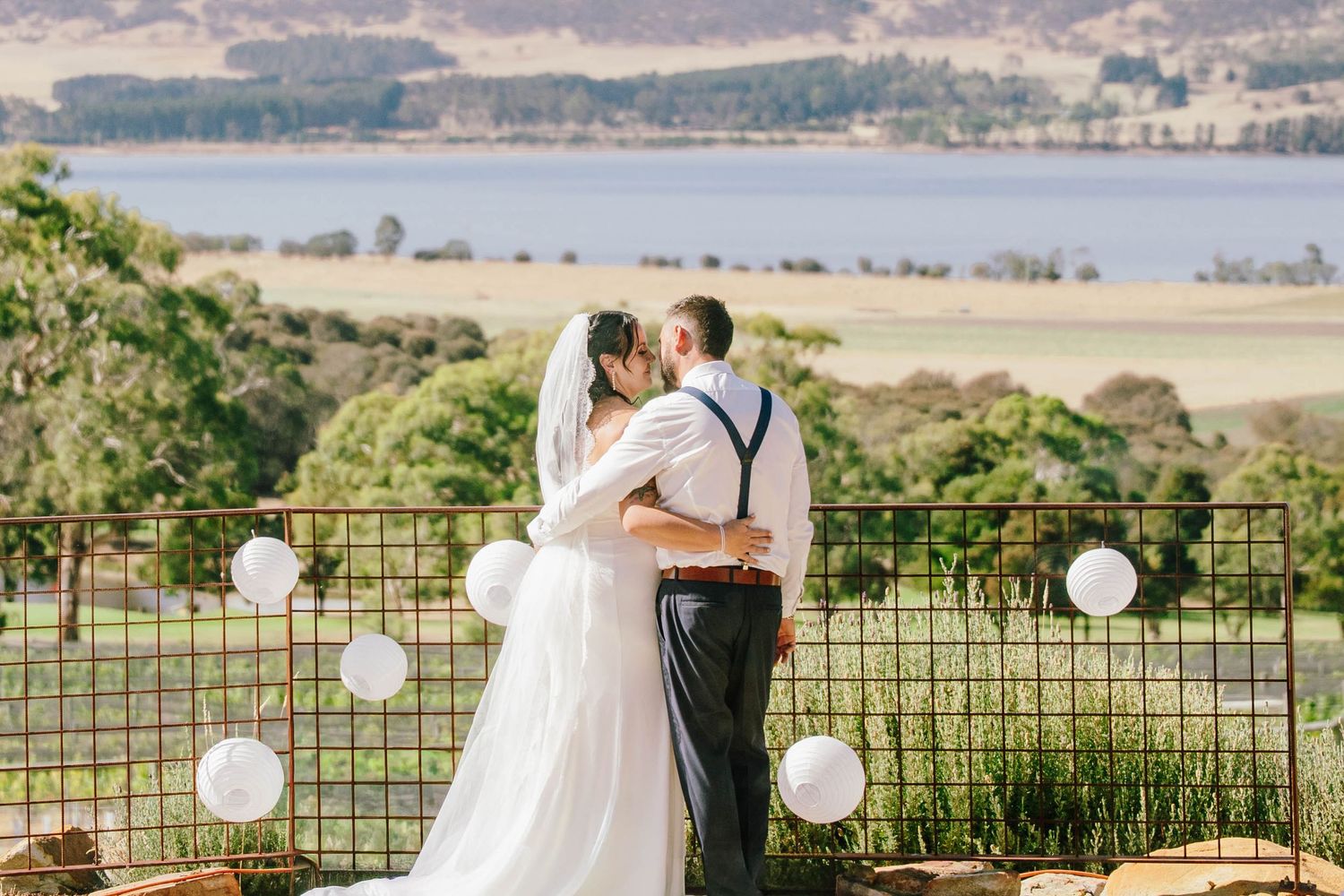 Bride and groom sharing a kiss at outdoor wedding ceremony with scenic lake and green hills backdrop.