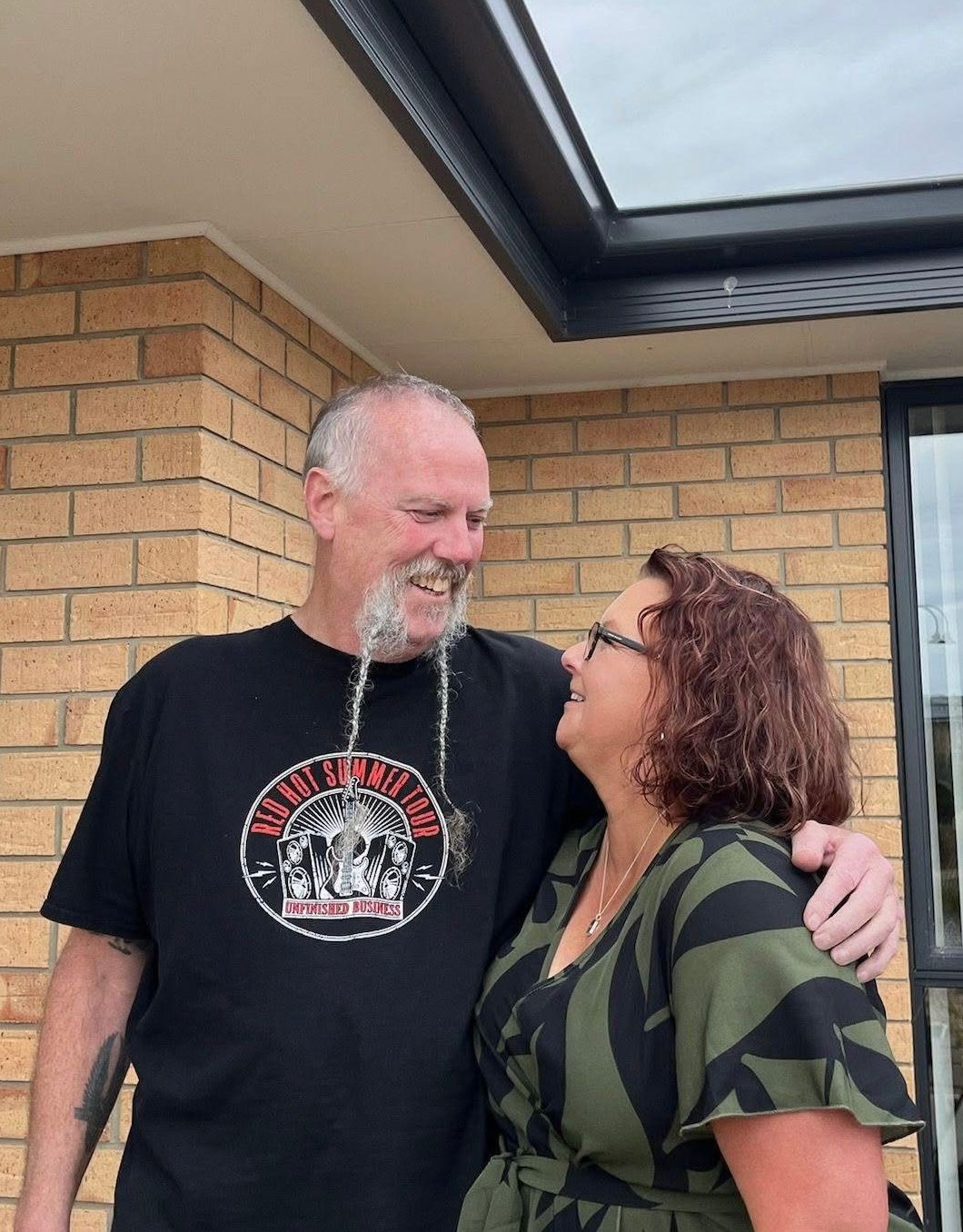 An older couple smiling and embracing outside a brick building, man wearing a black graphic t-shirt.