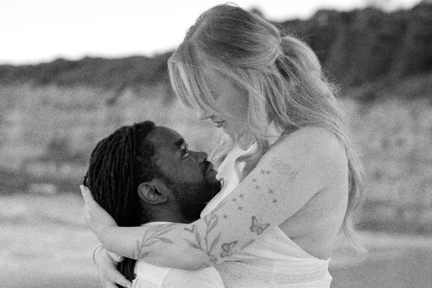Black and white romantic photo of a couple embracing and about to kiss outdoors, woman with blonde hair and man with dreadlocks.