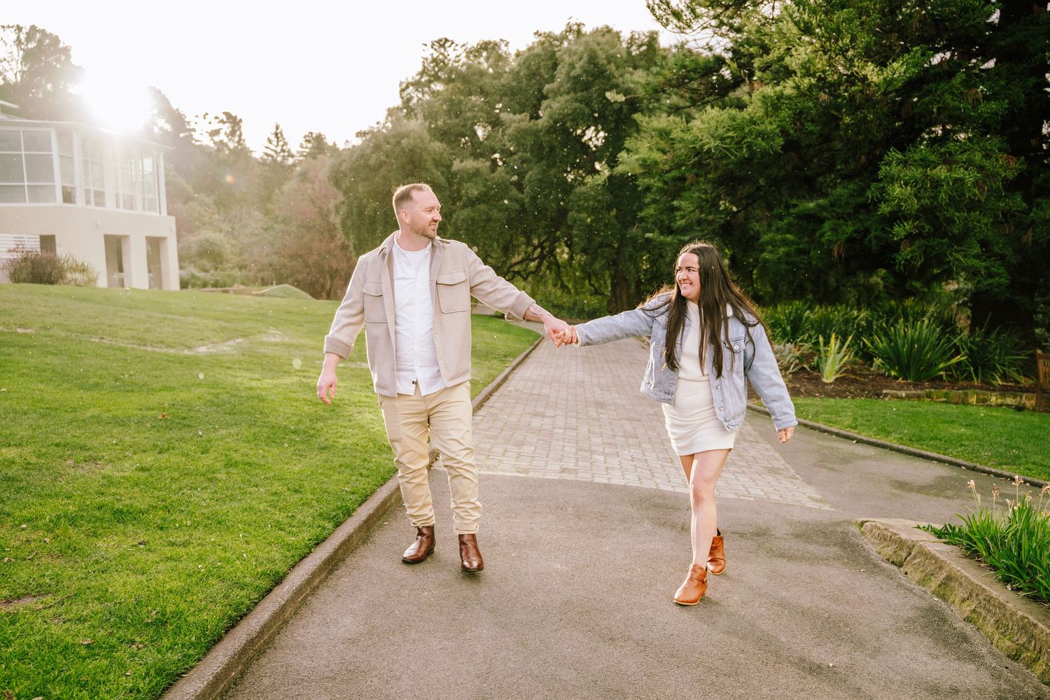 A happy couple walks hand-in-hand along a garden path, smiling at each other in warm golden sunlight.