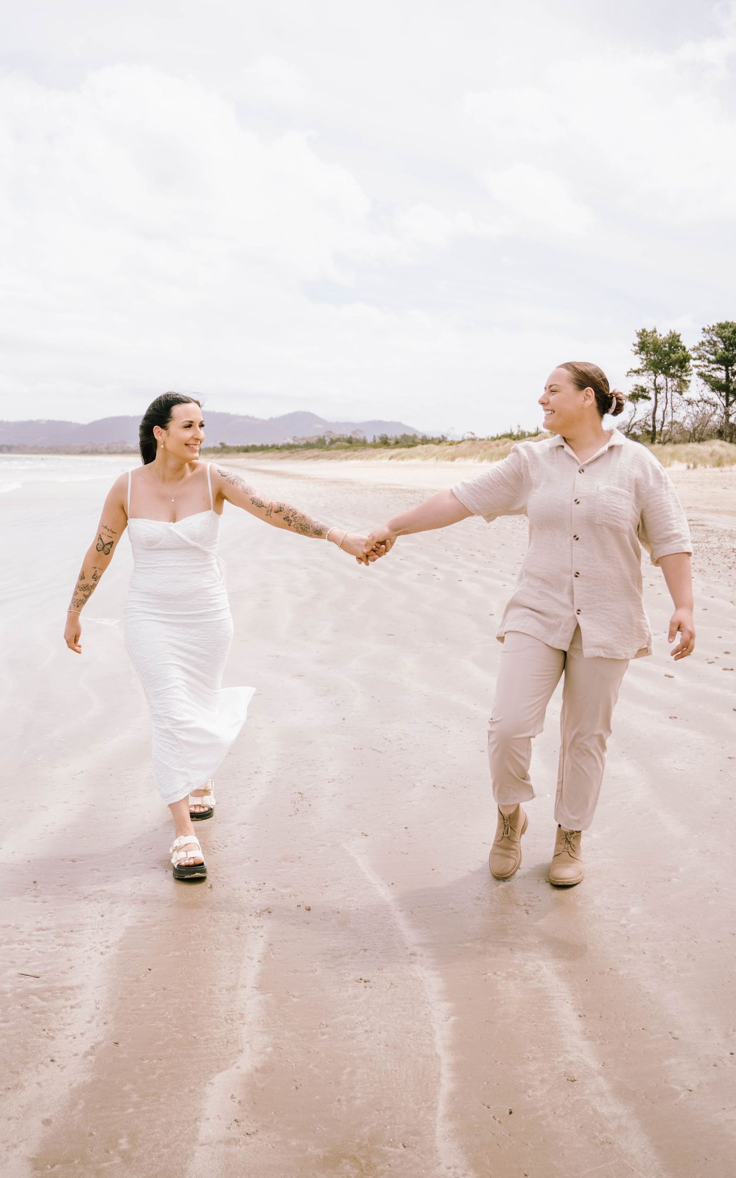 Playful couple holding hands and dancing together on a sandy beach with mountains in background.