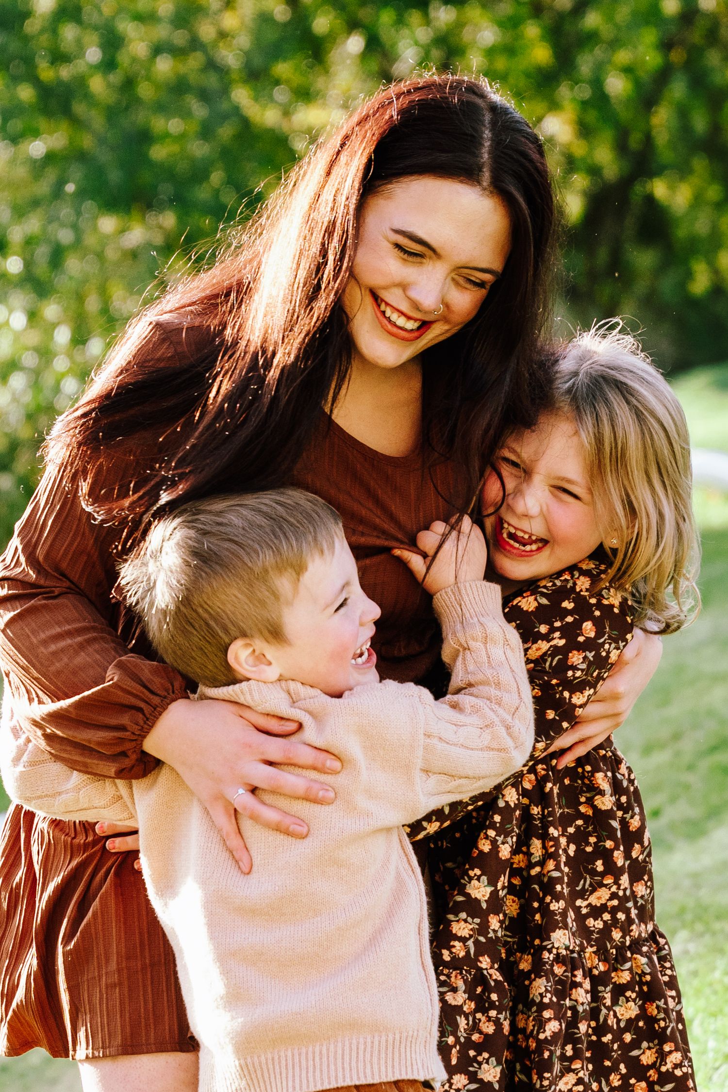 A smiling young woman hugs two laughing children outdoors in a sunlit green park setting.