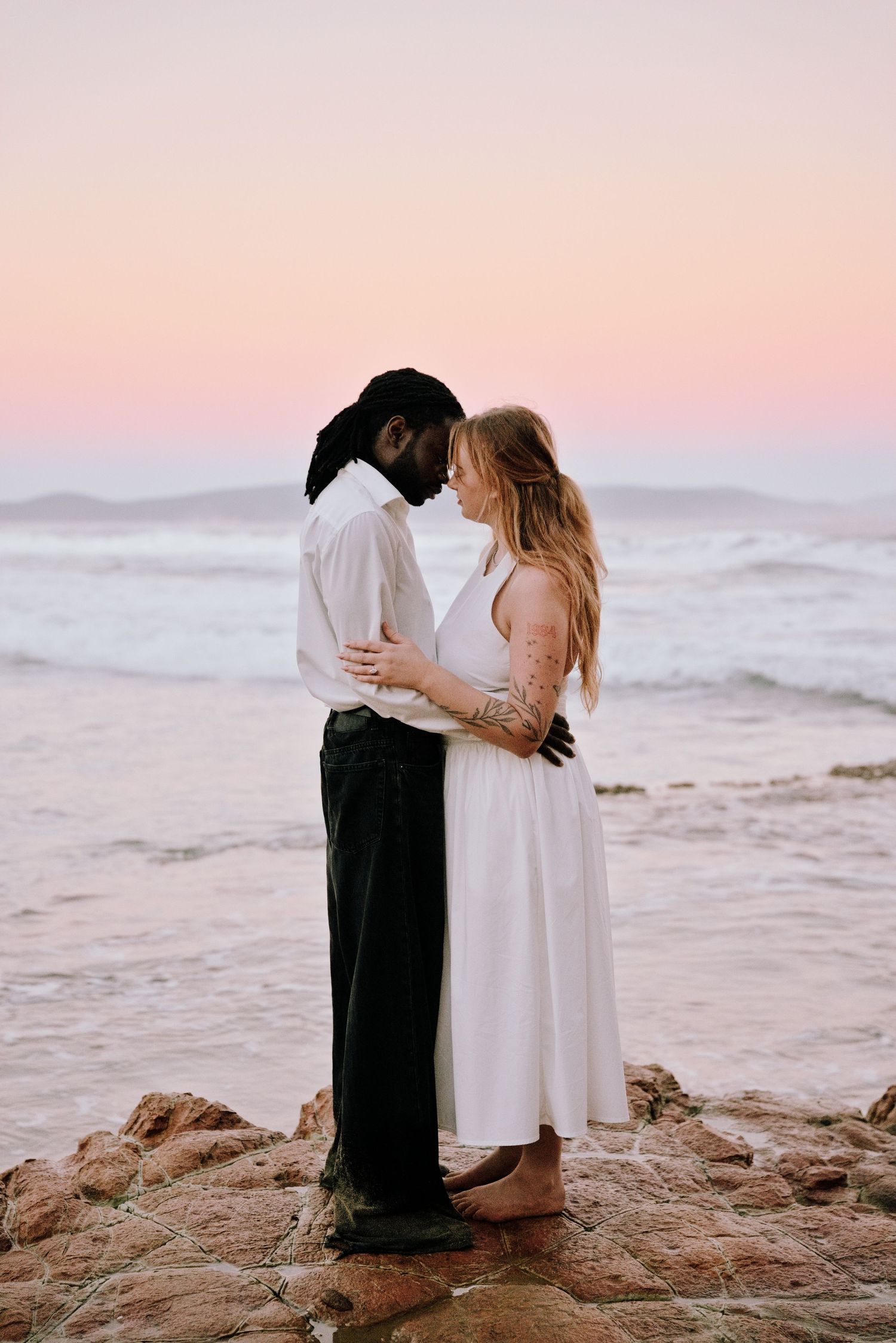 Couple embracing on rocky beach at sunset, woman in white dress and man in white shirt and black pants, ocean waves behind them.