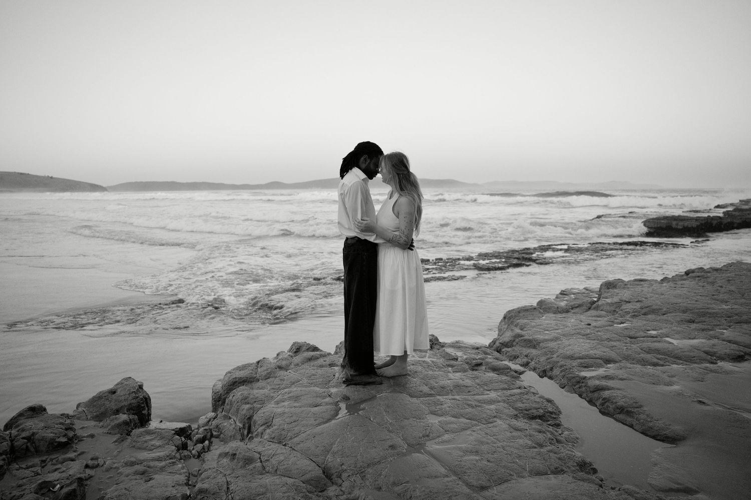 Black and white photo of a couple embracing on rocky shore with ocean waves behind them at dusk.