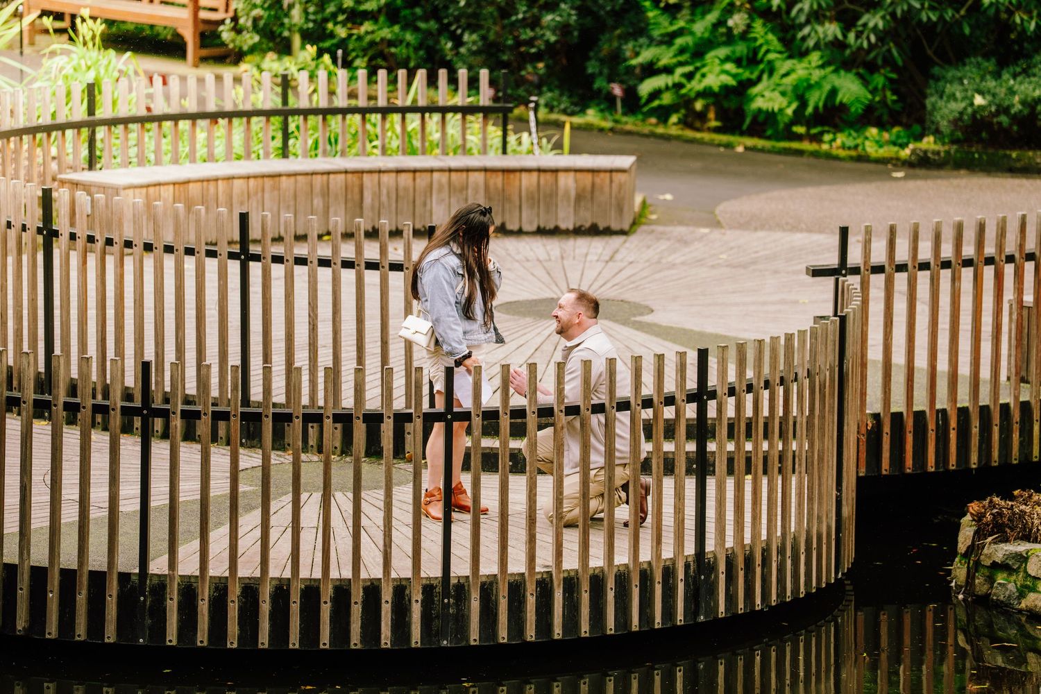Couple sharing a romantic moment on a curved wooden bridge over a pond in a lush green park setting.