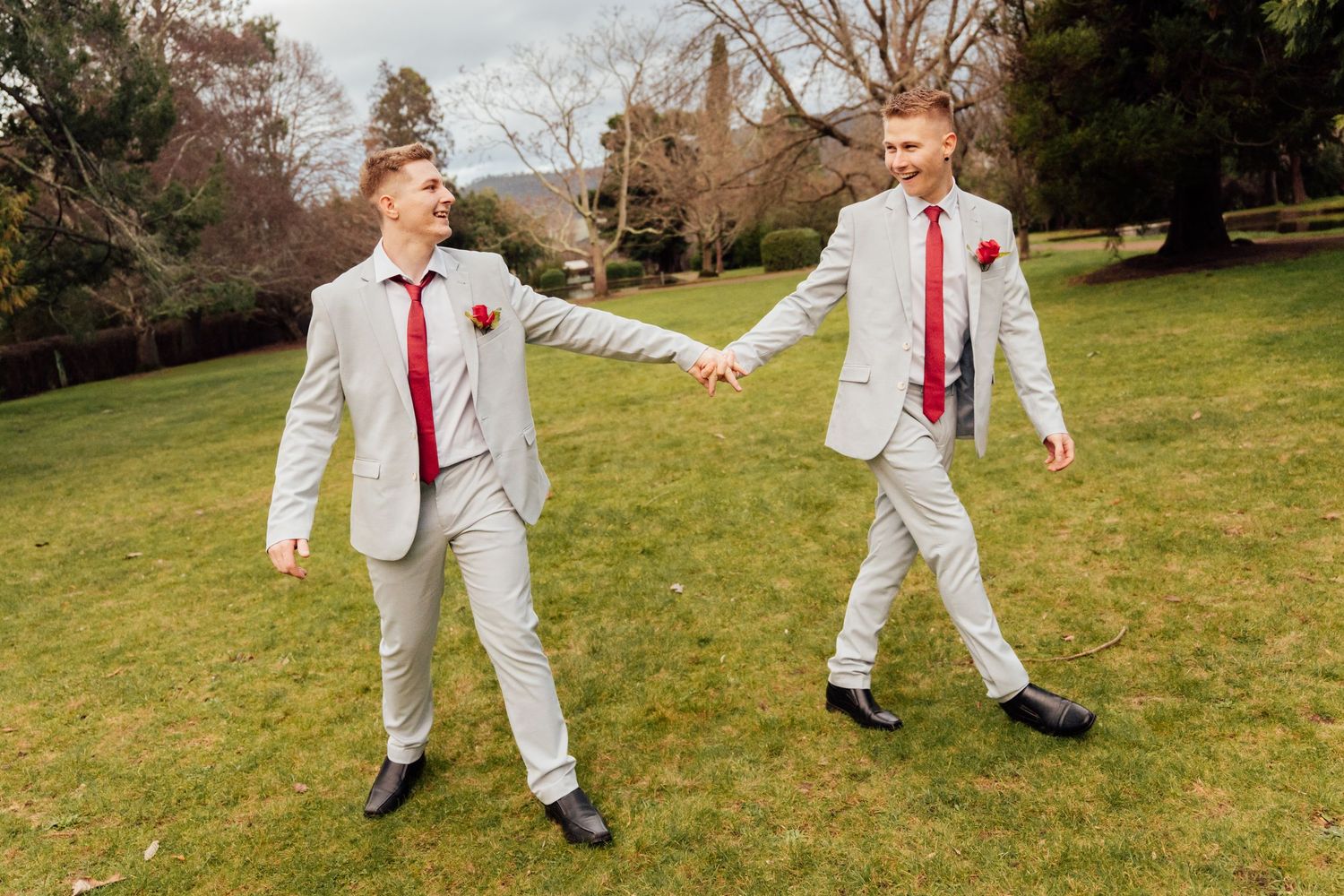 Two grooms in matching grey suits and red ties hold hands smiling on a green lawn.