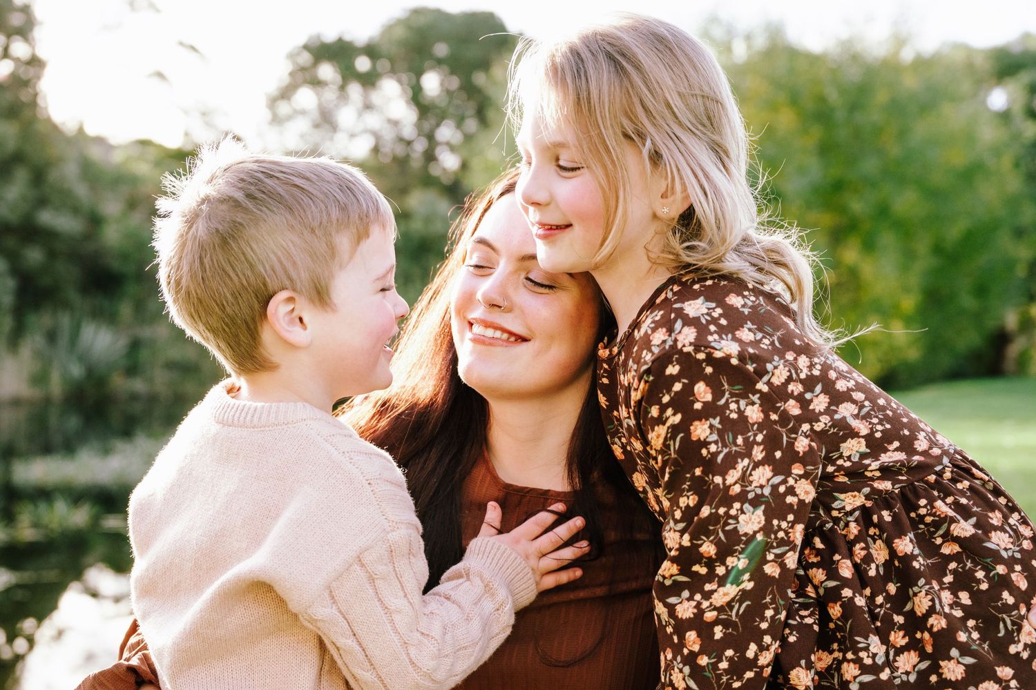 A smiling mother embraces her two blonde children outdoors in a warm, sunlit garden setting.