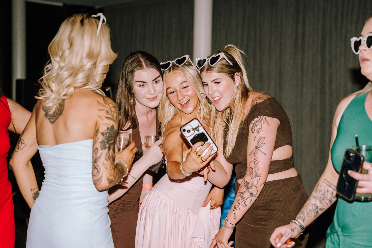 Group of tattooed women in party dresses taking a selfie together at an indoor social event.